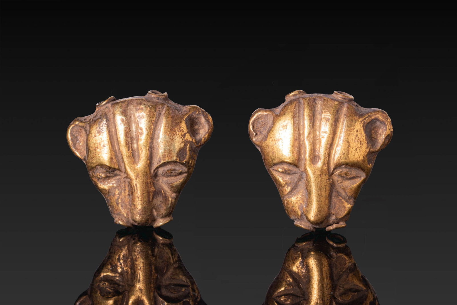 BEAUTIFUL PAIR OF GREEK GOLD LION-SHAPED APPLIQUES: Ca. 500 BC. A pair of double-sided gold lion head appliques, each with projecting rounded ears, downcast almond-shaped eyes, a tapering muzzle, and grooves on the forehead, pierced lengthwise with two
