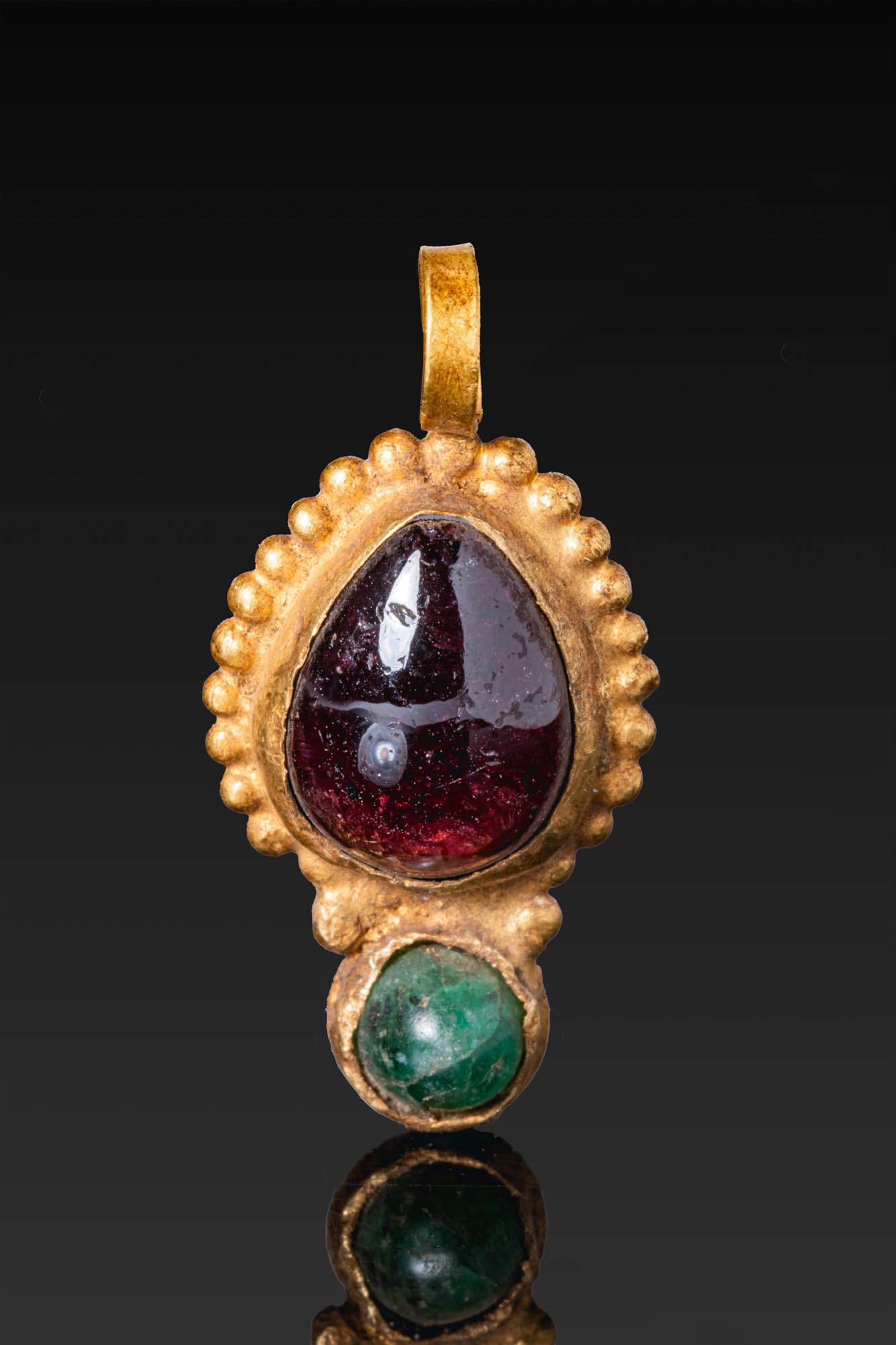 GREEK GOLD PENDANT WITH LARGE GARNET CABOCHON AND EMERALD: Ca. 400 - 200 BC. A gold pendant set with a large drop-shaped garnet cabochon within a beaded frame, with a smaller circular emerald below and a suspension loop at the apex. Size: 27mm x 13mm; Weight: