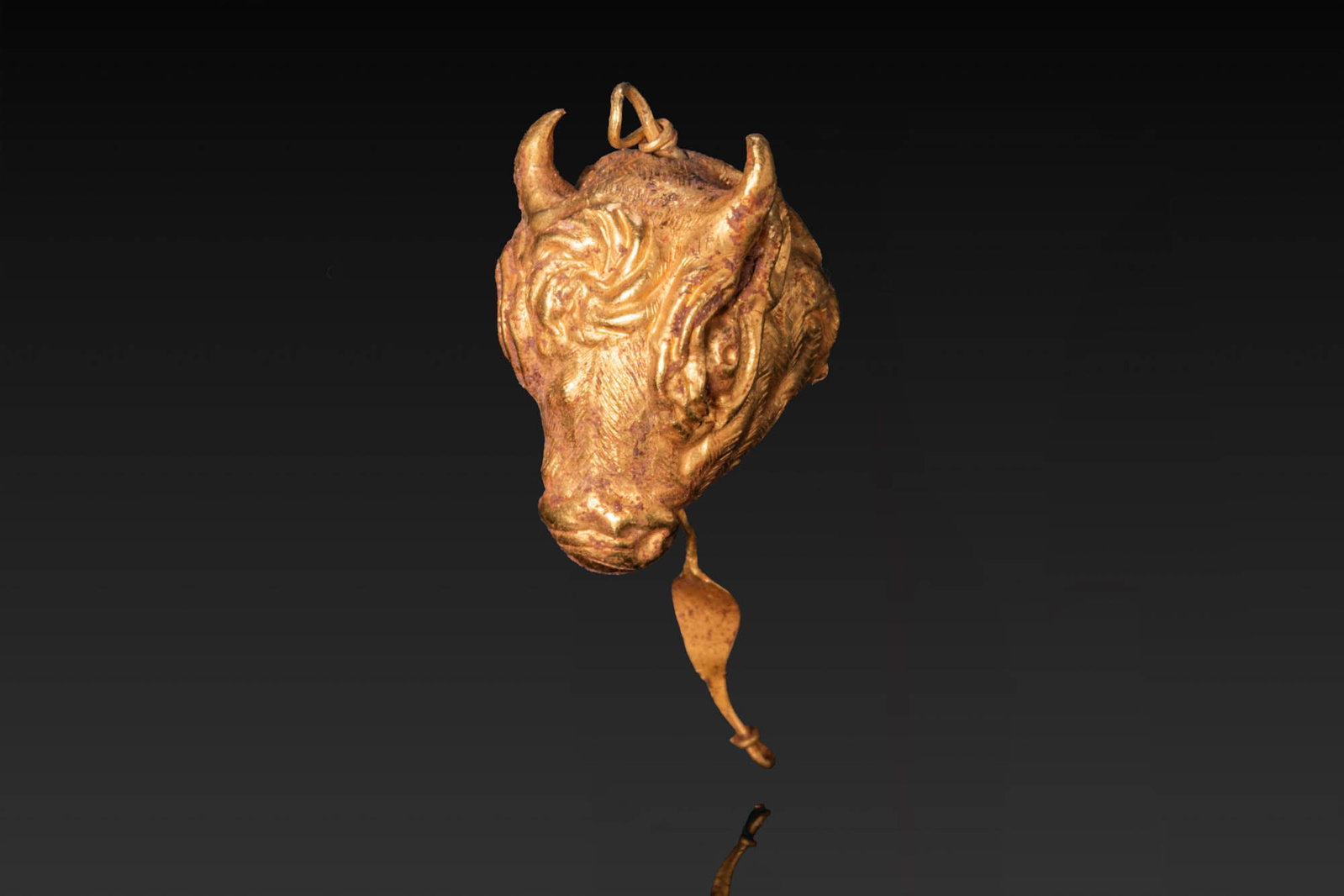 RARE GREEK PERIOD GOLD BULL'S HEAD PENDANT: Ca. 500 BC. A gold bull's head pendant with curved horns, large eyes, a tapering muzzle, and a curling forelock. A suspension loop is set above, and a leaf-shaped gold pendant hangs below. Size: 30mm 