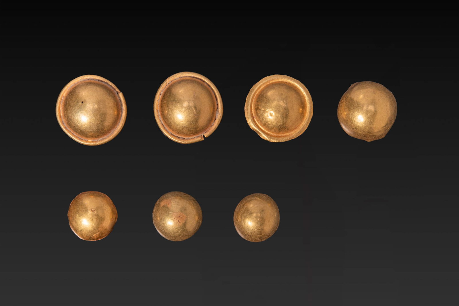 COLLECTION OF GREEK HIGH KARAT GOLD APPLIQUES: Ca. 800 - 600 BC. A collection of seven gold appliques, each formed as a domed disc of thin sheet with a raised or plain rim and a soldered attachment loop on the reverse. Size: 8-11mm x 8-11mm; Weigh