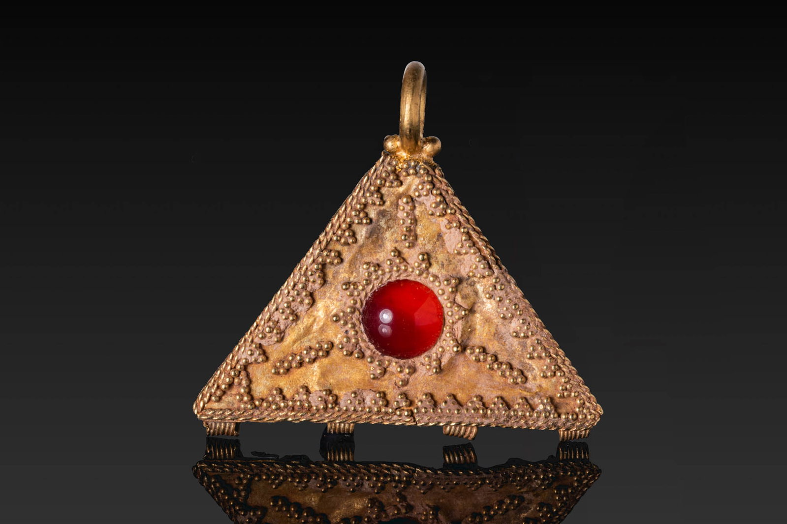 HELLENISTIC GOLD TRIANGULAR PENDANT WITH RED CABOCHON: Ca. 300 - 100 BC. A gold triangular pendant centred on a red cabochon in a circular mount. The obverse is decorated with fine granulation, arranged in a beaded outer border and in symmetrical clusters