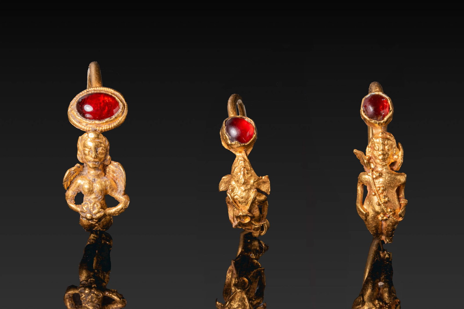 COLLECTION OF HELLENISTIC GOLD EARRINGS WITH EROS AND GARNETS: Ca. 200 - 100 BC. A collection of three gold earrings, each with a tapering hoop, one end narrowing to a point, and the other finished with a standing winged Eros. The nude figures stand with their ha
