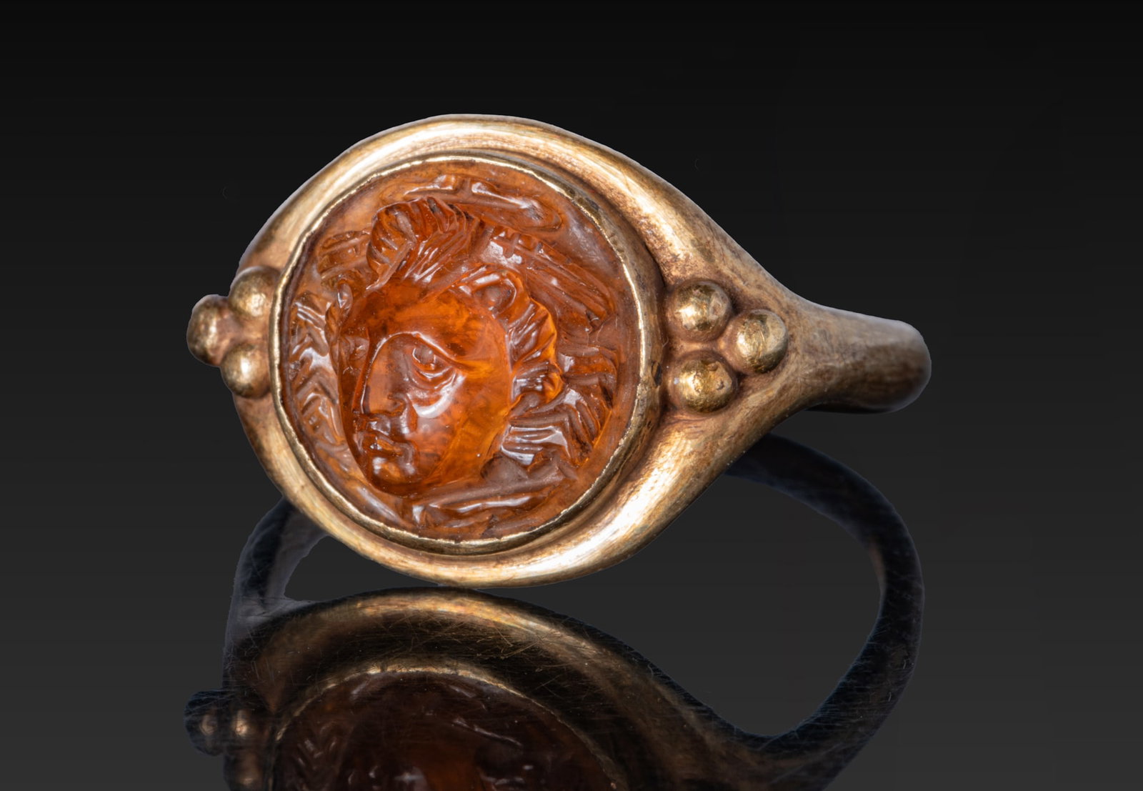 LATE ROMAN GOLD RING WITH STONE CAMEO OF A GORGON: Ca. AD 200 - 400. A gold ring with a rounded hoop expanding to an oval bezel with a plain collet set with an orange stone cameo carved with the head of a Gorgon, her gaze lowered, with snakes radiatin