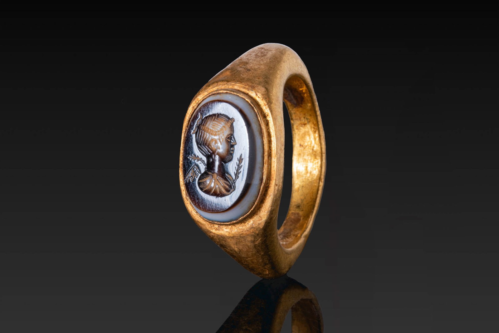 LARGE ROMAN GOLD RING WITH AGATE INTAGLIO OF CERES: Ca. AD 200 - 300. A gold ring with a broad, rounded hoop and angular shoulders leading to an oval bezel set with a layered agate intaglio engraved with the right-facing bust of Ceres, her hair bound w