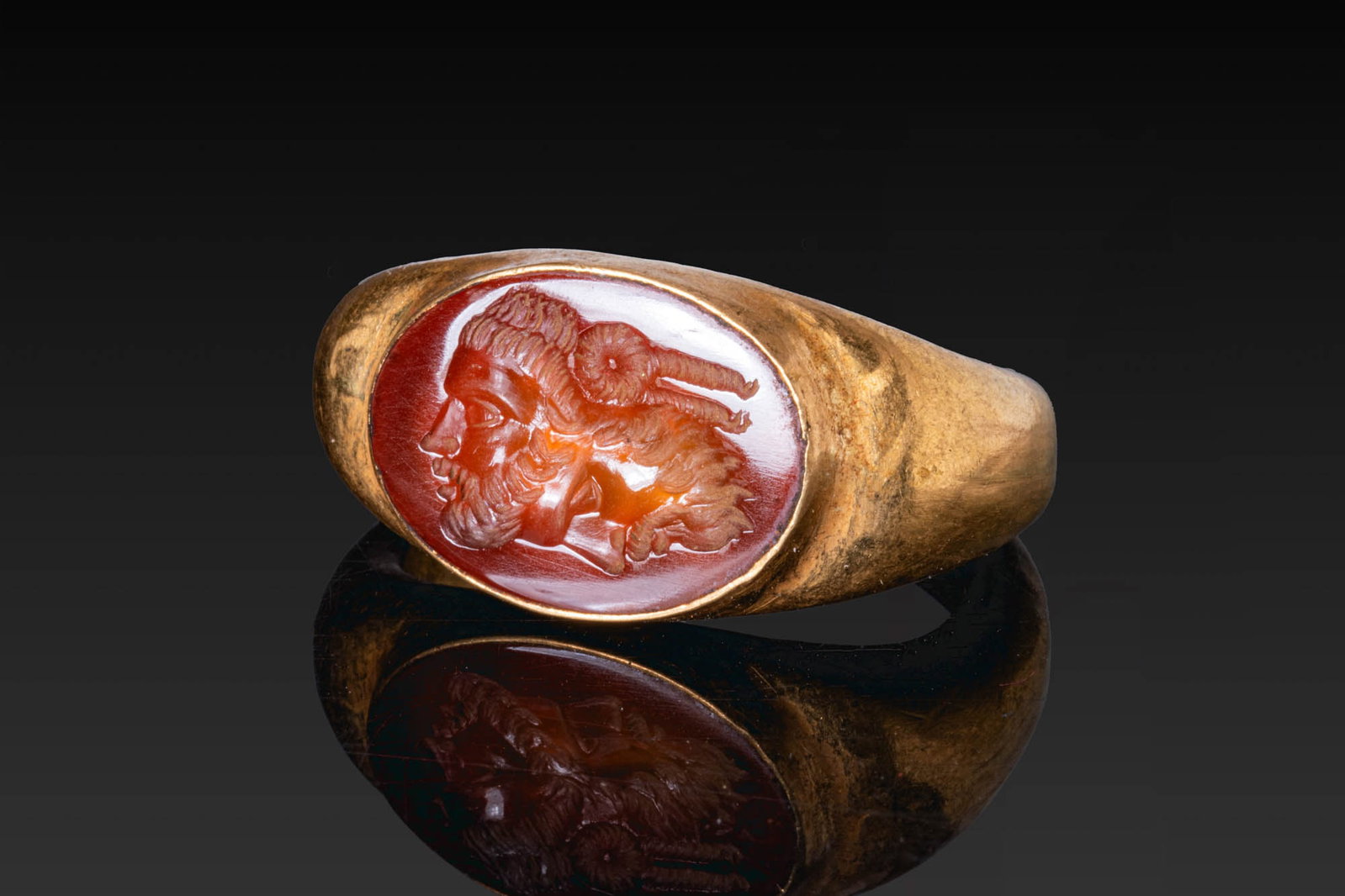 ROMAN CARNELIAN INTAGLIO OF A GRYLLOS IN GOLD RING: Ca. AD 1 - 300. A carnelian intaglio engraved with a gryllos composed of a bearded male head in left profile conjoined at a right angle with a second bearded head turned downward. It is set in a possi