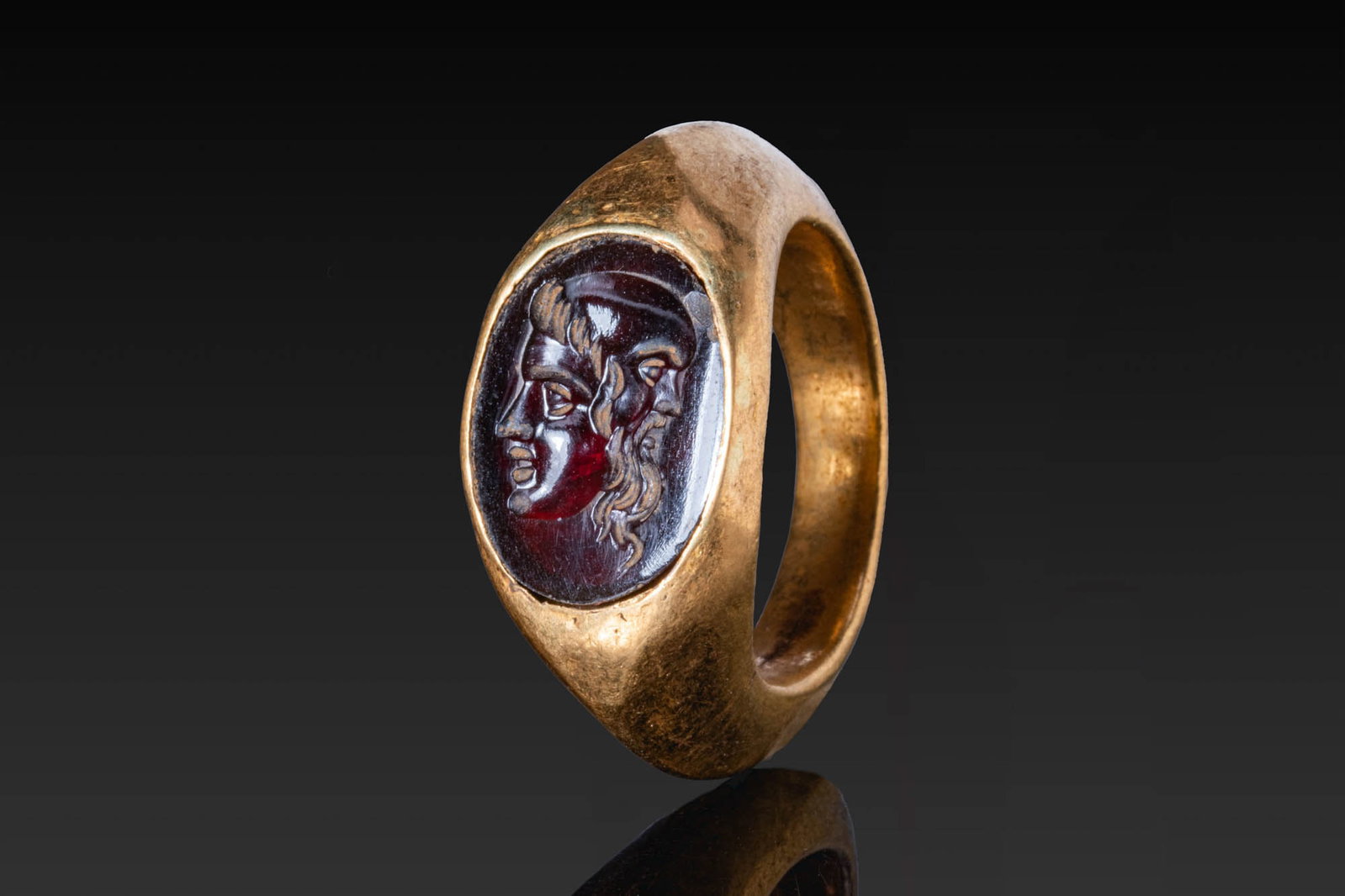 ROMAN GARNET INTAGLIO OF A GRYLLOS IN GOLD RING: Ca. AD 1 - 300. A garnet intaglio engraved with a janiform gryllos composed of two conjoined faces, a youth looking to the left and a bearded bald Silenus to the right. It is set in the oval bezel of 