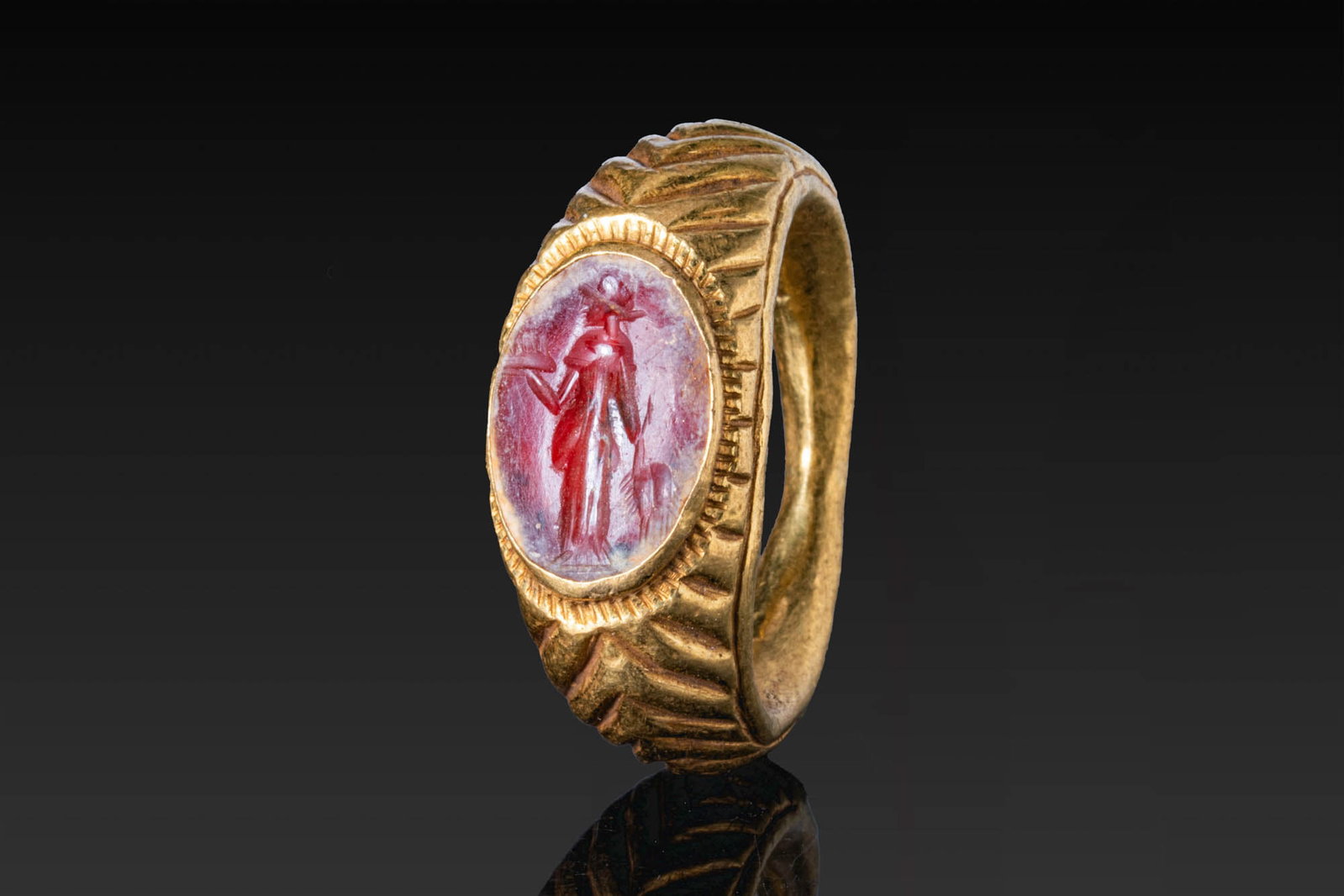 ROMAN CARNELIAN INTAGLIO OF MINERVA IN GOLD RING: Ca. AD 50 - 200. A carnelian intaglio engraved with Minerva standing in profile, wearing a helmet and long drapery, mounted in a possibly later gold ring with an expanding hoop, chevroned shoulders, a