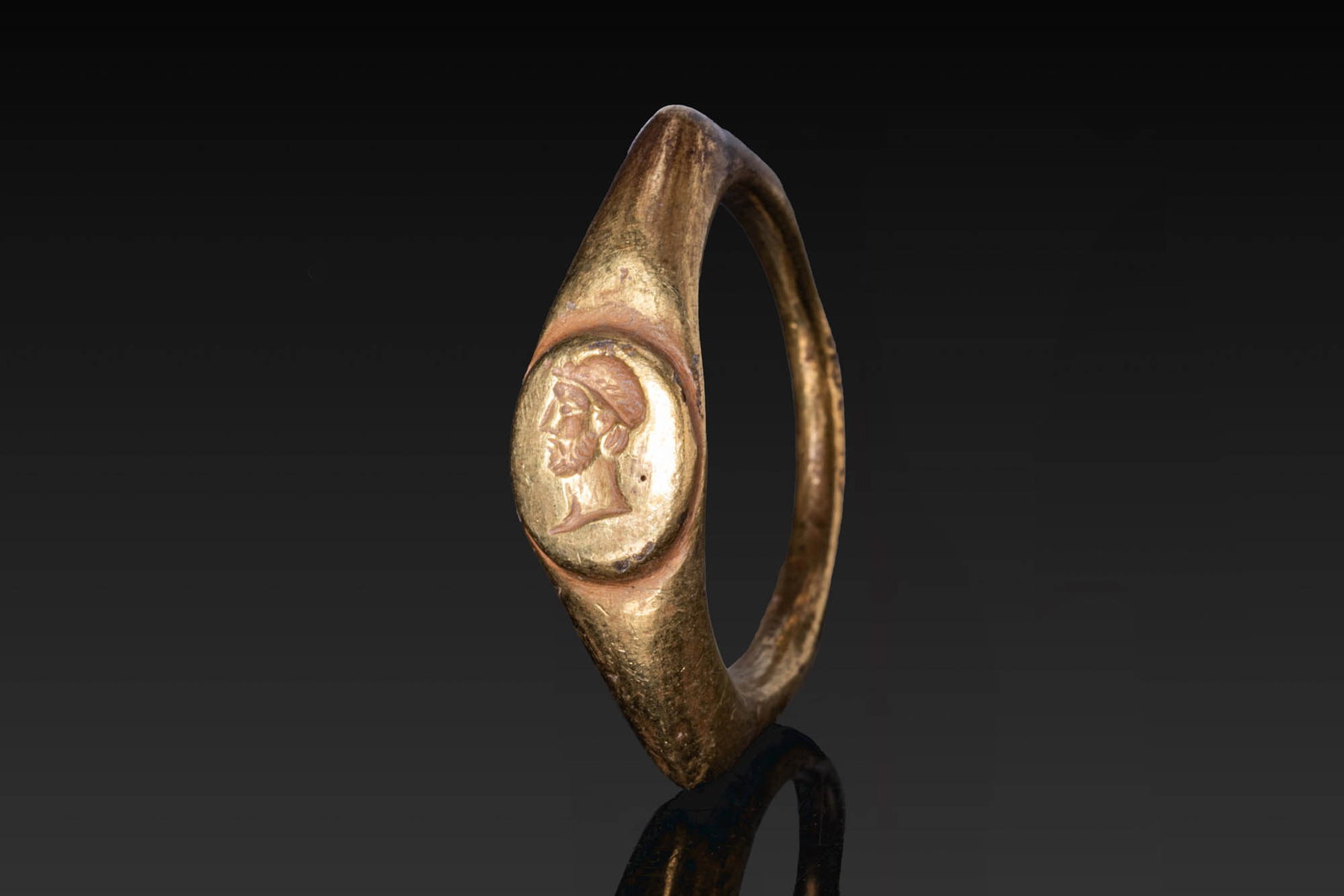 ROMAN GOLD RING WITH PROFILE PORTRAIT OF EMPEROR: Ca. AD 200 - 300. A gold ring with a slender hoop, angular shoulders, and a raised oval bezel engraved with a portrait of an emperor, possibly Gallienus, in left profile, wearing a close-fitting cap. 