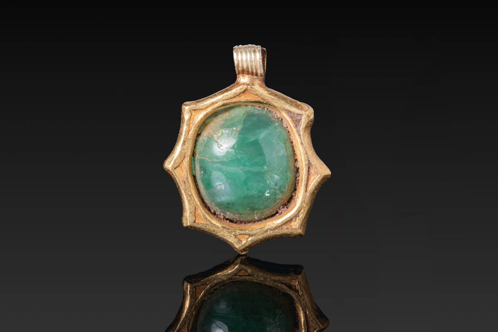 LATE ROMAN GOLD PENDANT WITH LARGE EMERALD CABOCHON ENGRAVED ON THE UNDERSIDE WITH A PROFILE PORTRAI: Ca. AD 300 - 600. A gold pendant with a star-shaped frame centred by an oval emerald. The reverse is engraved with the head of Hercules in profile, bearded and with curling hair. A ribbed suspension l