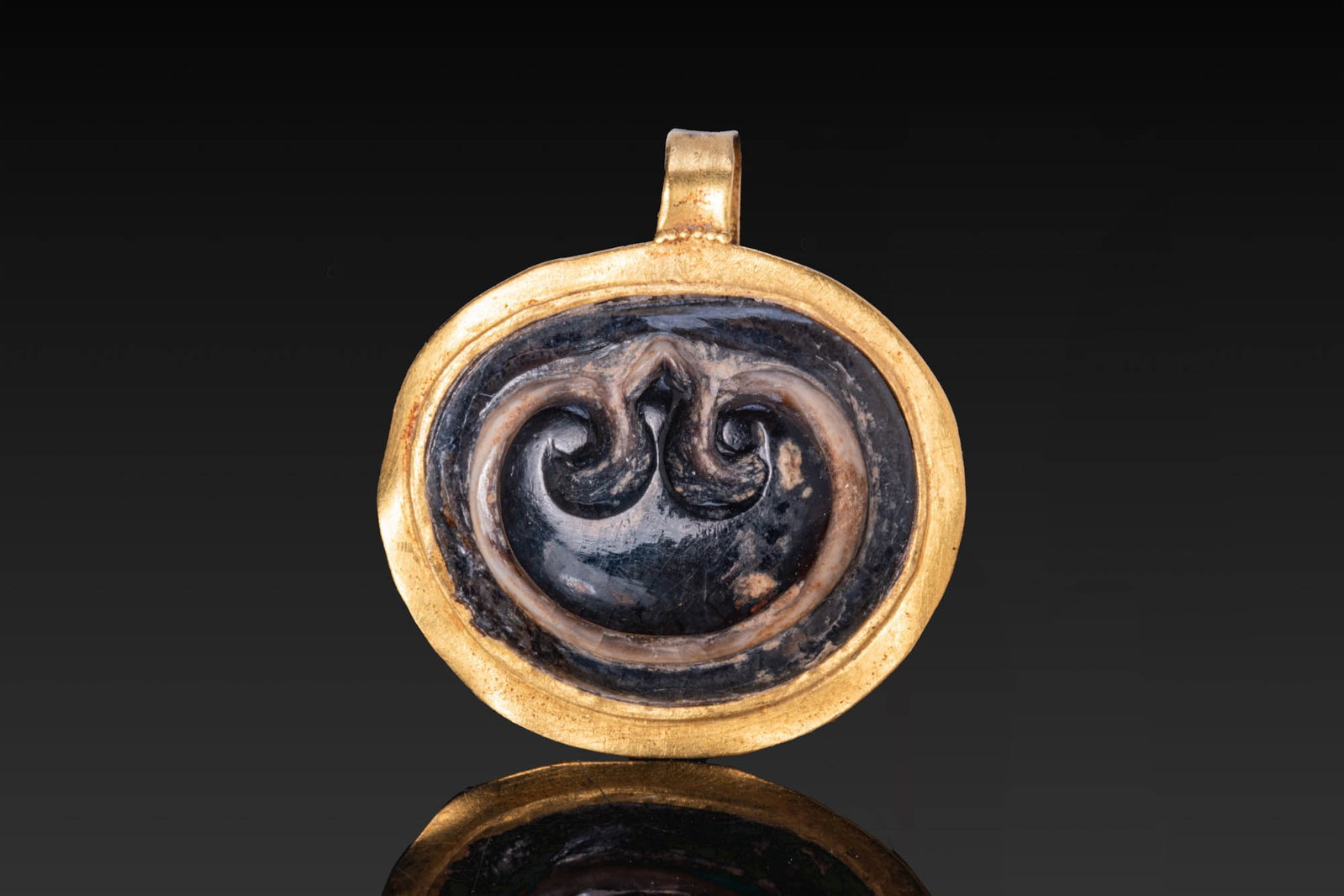 ROMAN GOLD PENDANT WITH AGATE CAMEO OF A PELTA: Ca. AD 1 - 300. A gold pendant with an oval frame and a plain suspension loop, set with an agate cameo carved with a pelta, a crescent-shaped shield with inward curving ends. Size: 21mm x 19mm; Weight