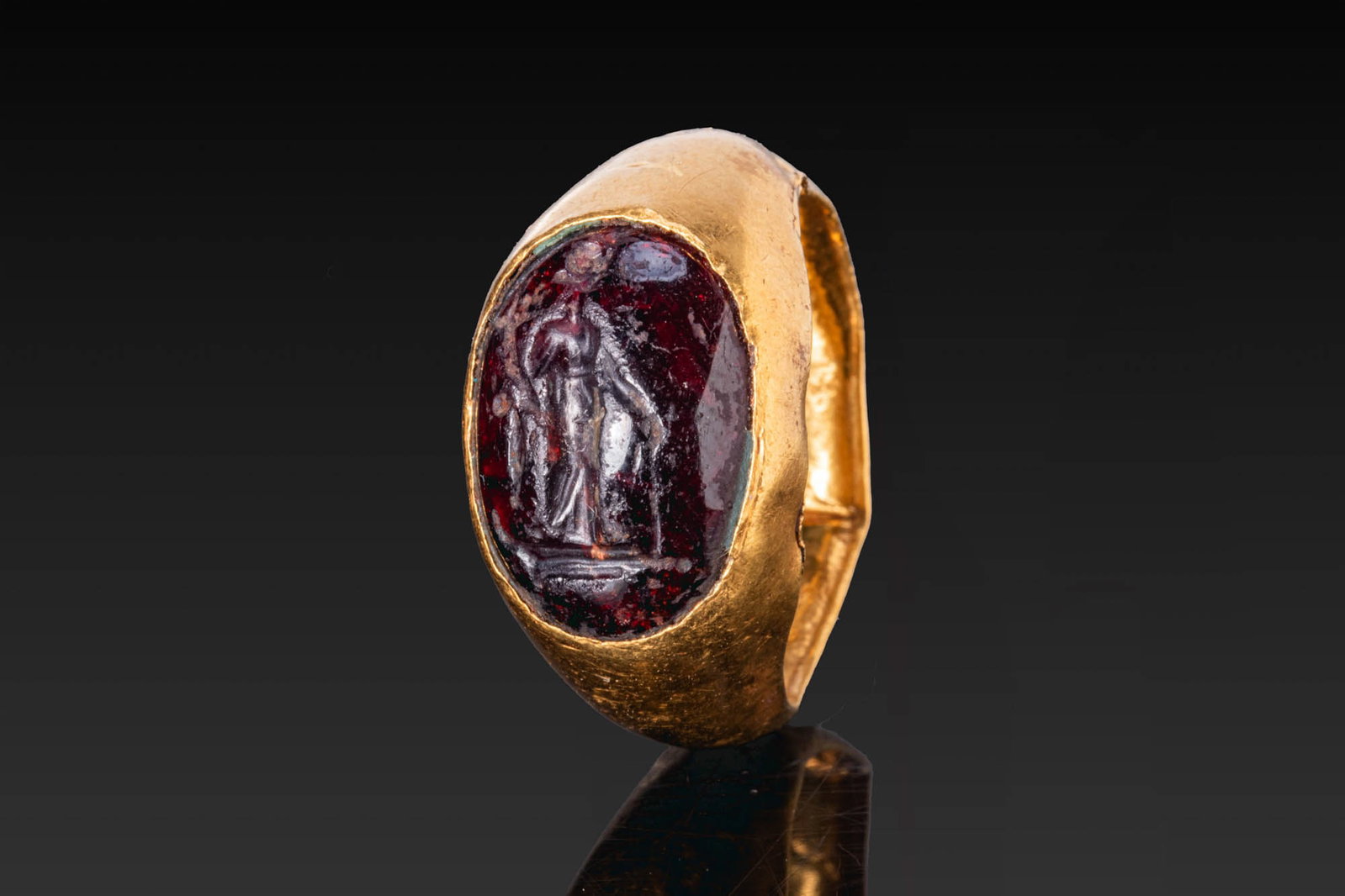 LATE ROMAN GOLD RING WITH RED GARNET INTAGLIO OF FORTUNA: Ca. AD 100 - 300. A gold hollow ring with a tapering hoop and an oval bezel set with a convex red garnet intaglio engraved with Fortuna standing in three-quarter view, holding a cornucopia in her righ