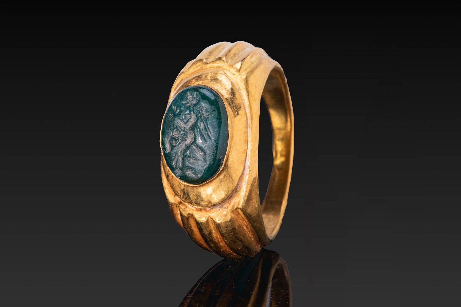 ROMAN GOLD RING WITH GREEN STONE GEM OF CUPID: Ca. AD 300. A gold ring with a slender hoop, fluted shoulders, and a raised oval bezel cell set with a green stone gem engraved with winged Cupid walking to the left. For similar see: The Classical Ar