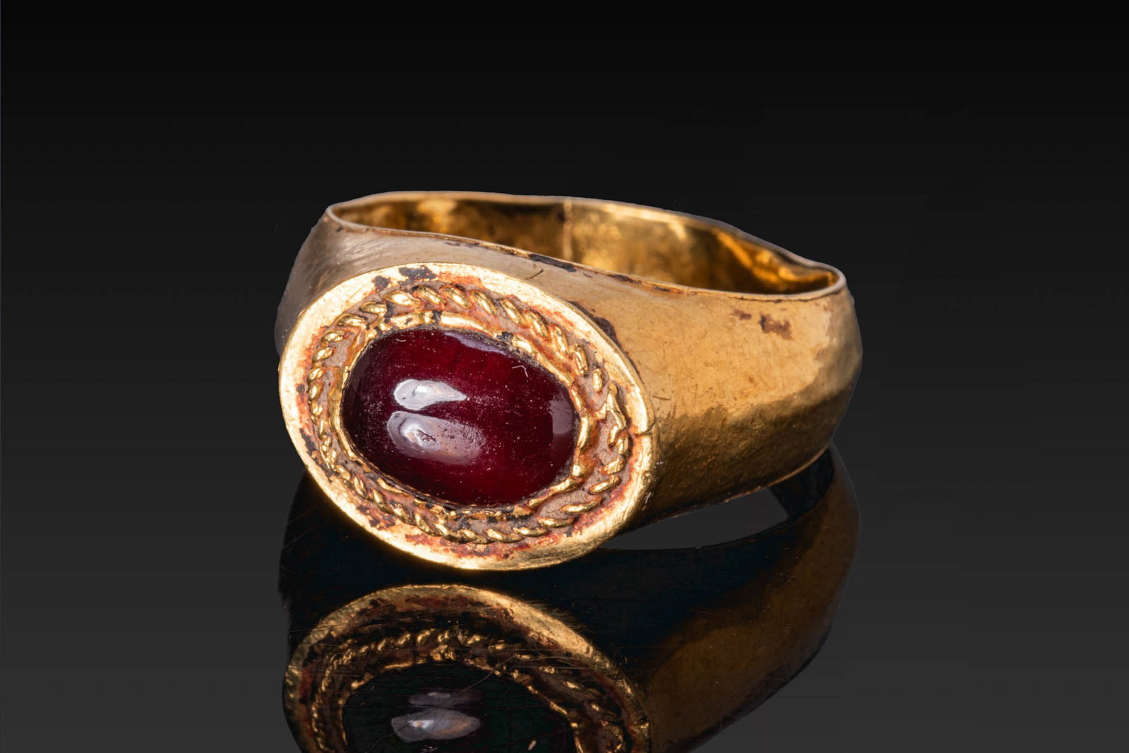 ROMAN GOLD RING WITH CENTRAL RED GARNET CABOCHON: Ca. AD 100 - 300. A gold ring with a broad tapering hoop and an oval bezel set with a red garnet cabochon within a twisted border. For similar see: The British Museum, Museum number 1917,0501.746. Siz