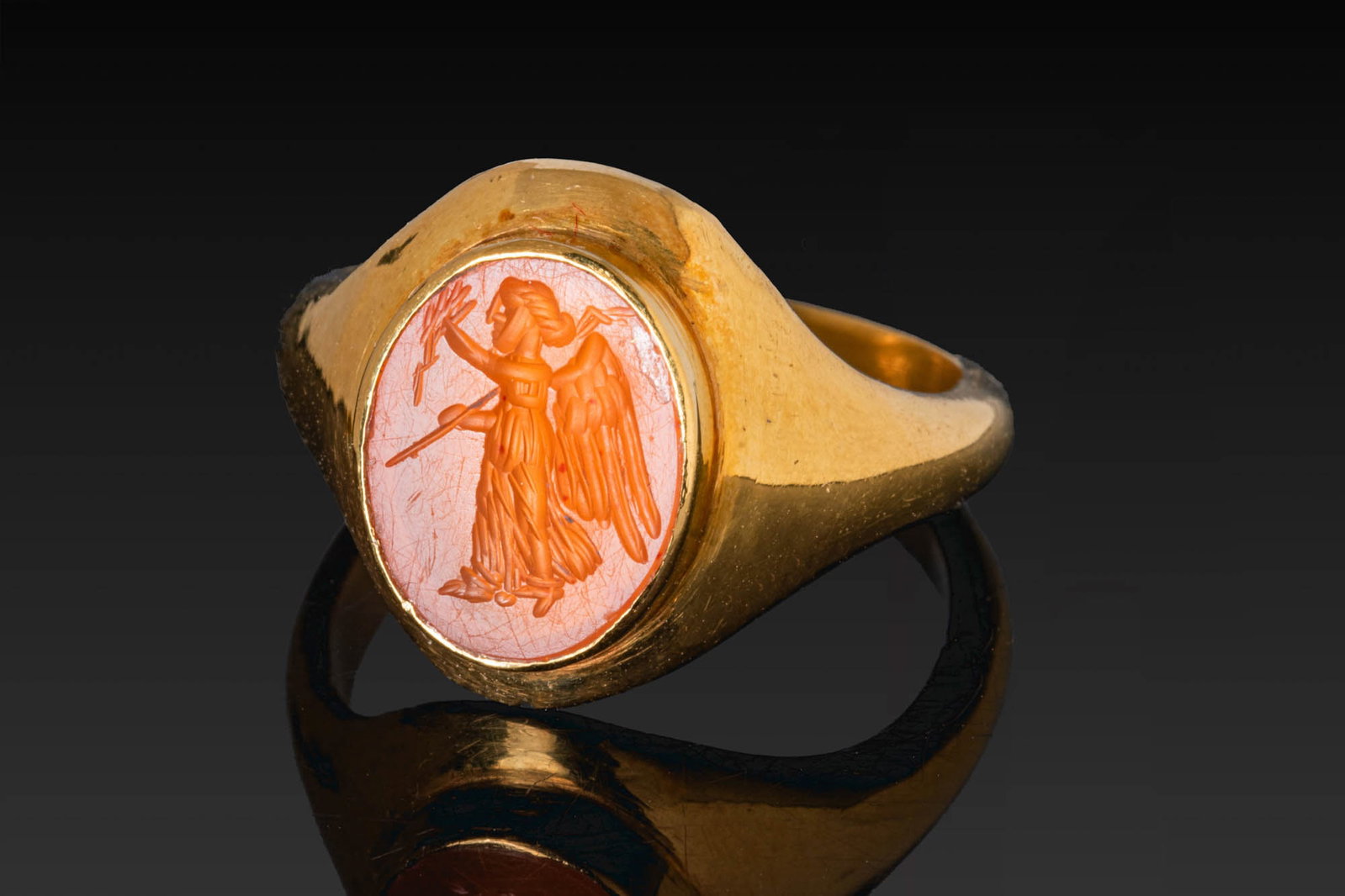 ROMAN CARNELIAN INTAGLIO OF VICTORY IN GOLD RING: Ca. AD 50 - 200. A carnelian intaglio engraved with winged Victory standing in profile, her left arm extended with a laurel wreath and a palm branch resting against her right shoulder, mounted in a po