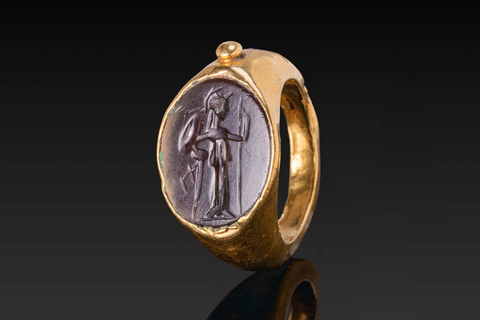 ROMAN GOLD RING WITH GARNET INTAGLIO OF MINERVA: Ca. AD 50 - 200. A gold hollow ring with a tapering hoop and an oval bezel set with a garnet intaglio engraved with Minerva standing in profile, helmeted, holding a shield and spear. A small applied b