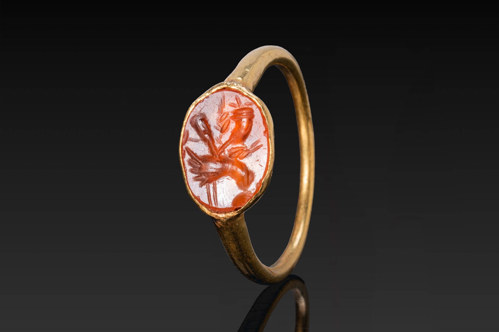 ROMAN CARNELIAN INTAGLIO OF A HAND HOLDING A CORNUCOPIA, EARS OF GRAIN AND A POPPY SEED HEAD IN GOLD: Ca. AD 200 - 400. A carnelian intaglio engraved with a hand holding a cornucopia, two ears of grain and a poppy seed head beside, mounted in a possibly later gold ring with a slender D-section hoop an