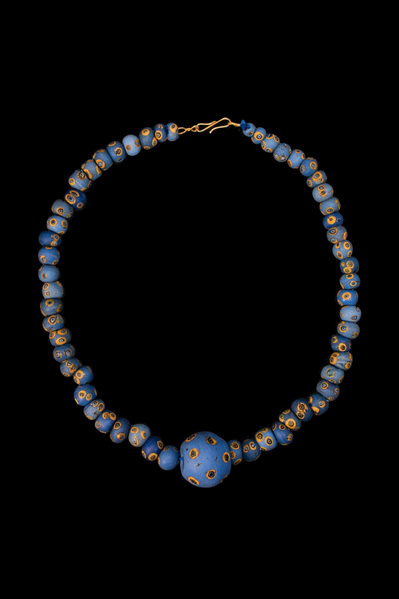 LARGE PHOENICIAN GLASS EYE BEAD NECKLACE: Ca. 600 - 300 BC. A necklace composed of blue glass beads of globular form, each decorated with yellow and black eye motifs, centred by a larger spherical bead and finished with a hook clasp. For simi