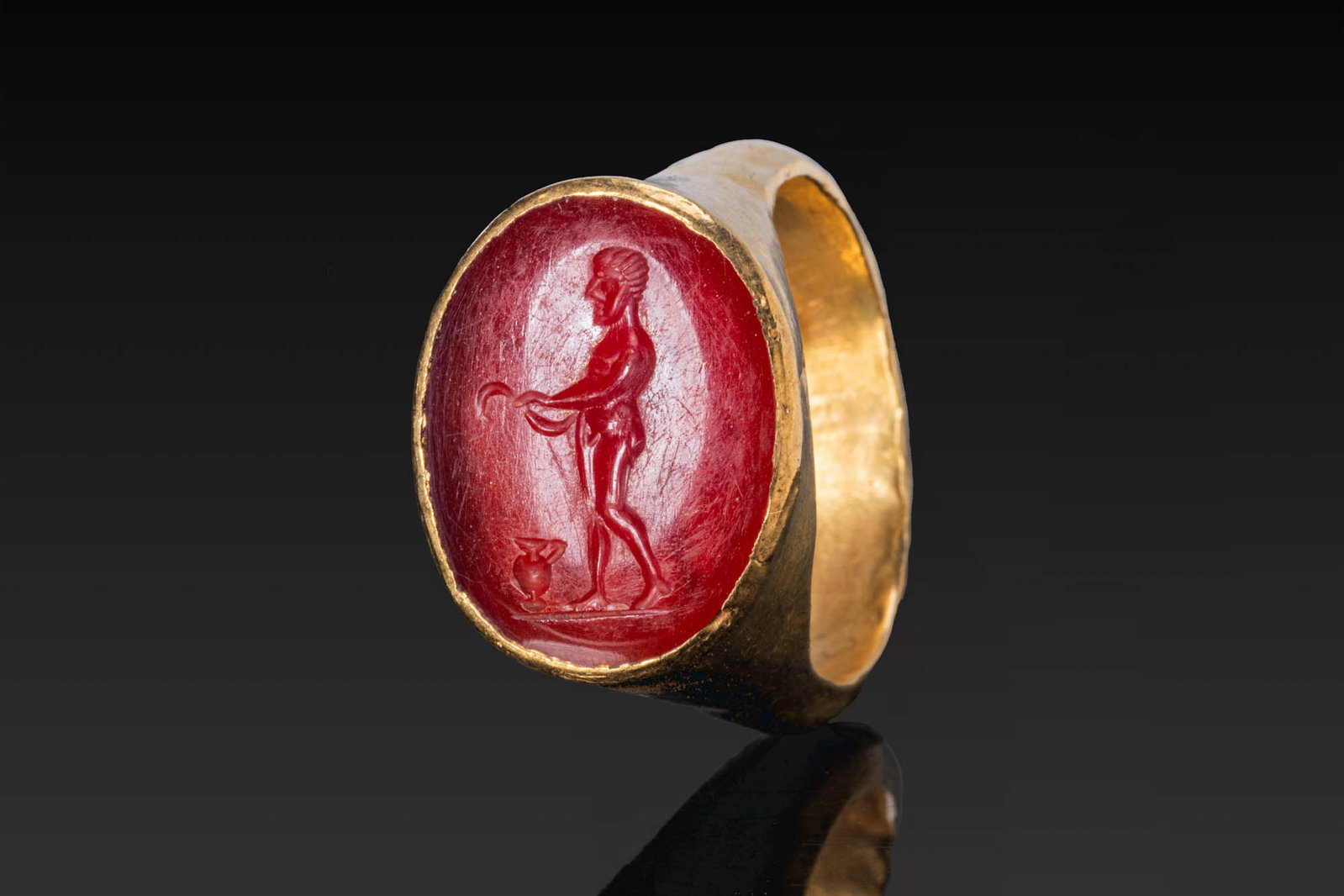 ROMAN GOLD RING WITH CARNELIAN INTAGLIO OF AN ATHLETE: Ca. AD 1 - 300. A gold ring with a tapering hoop and a tall oval bezel, set with a carnelian intaglio engraved with a nude athlete standing in profile on a groundline, with one arm extended and a wate