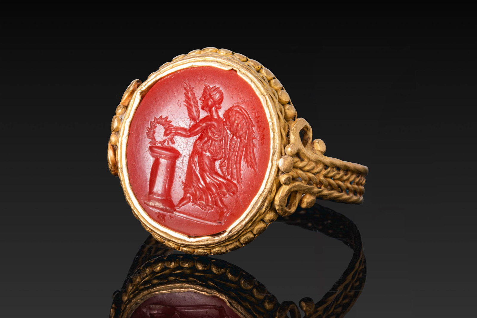 ROMAN CARNELIAN INTAGLIO OF WINGED VICTORY BEFORE AN ALTAR IN GOLD RING: Ca. AD 300. A carnelian intaglio engraved with winged Victory standing before an altar, extending a wreath in her left hand and holding a palm branch in her right, all on a groundline, mounted in a po