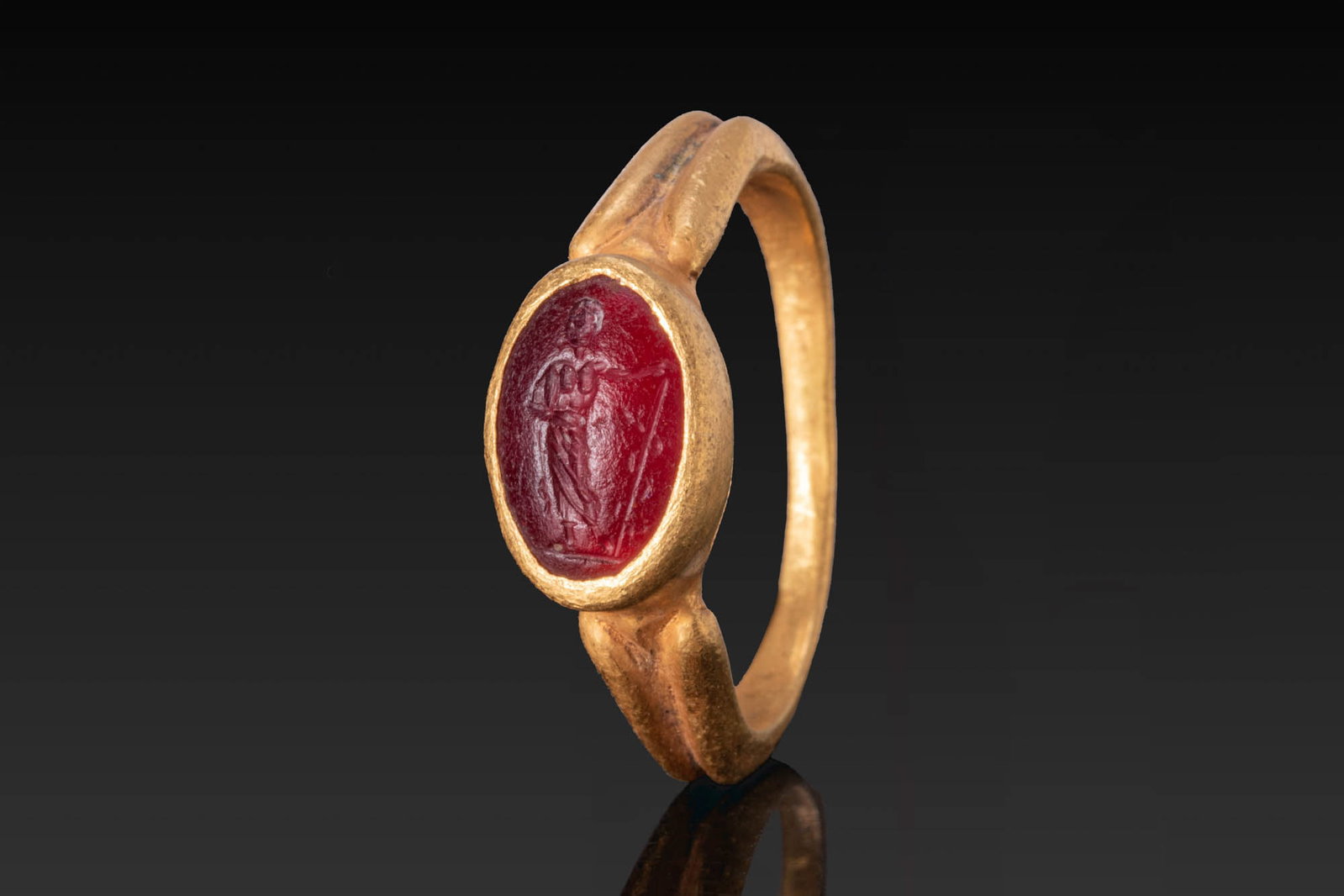 ROMAN GOLD RING WITH CARNELIAN INTAGLIO OF ASCLEPIUS: Ca. AD 300. A gold ring with a slender hoop broadening at the shoulders and an oval bezel set with a carnelian intaglio engraved with a draped figure of Asclepius standing to left and holding a staff,
