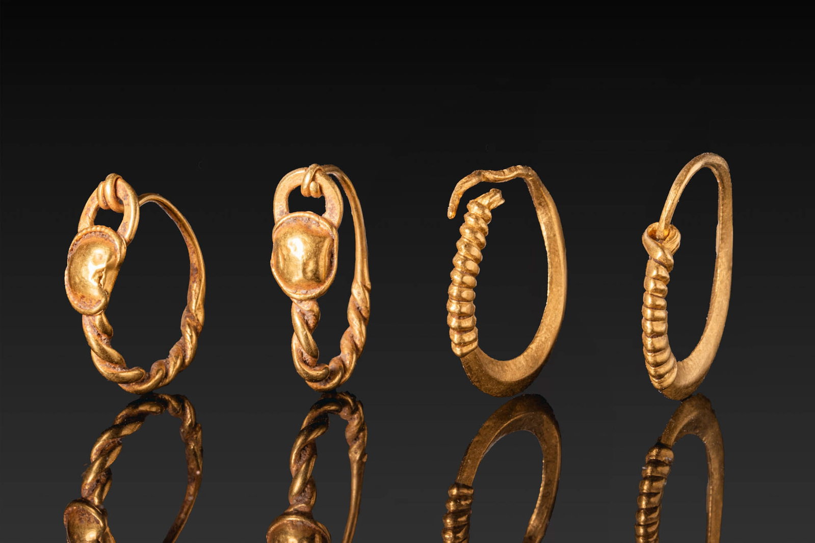 COLLECTION OF ROMAN GOLD EARRINGS: Ca. AD 1 - 300. A collection of four gold earrings, including, from left to right: a matched pair of hoop earrings with twisted lower sections and plain upper arcs, each centred on a circular boss in 