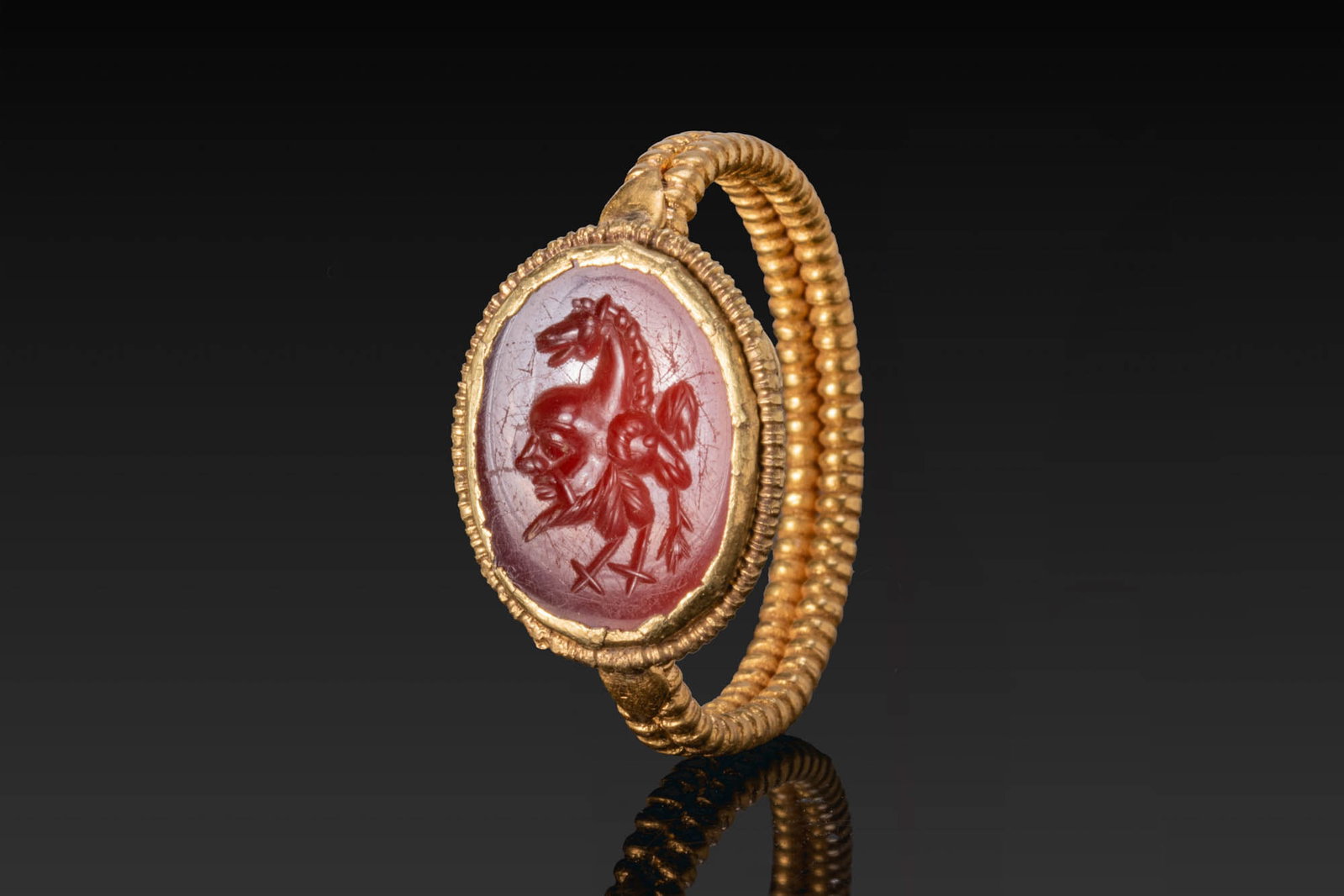 ROMAN CARNELIAN INTAGLIO OF A GRYLLOS IN GOLD RING: Ca. AD 300. A carnelian intaglio engraved with a gryllos composed of the conjoined heads of a Silenus, ram, and horse, on rooster legs, mounted in a possibly later gold ring with a ribbed hoop and an 