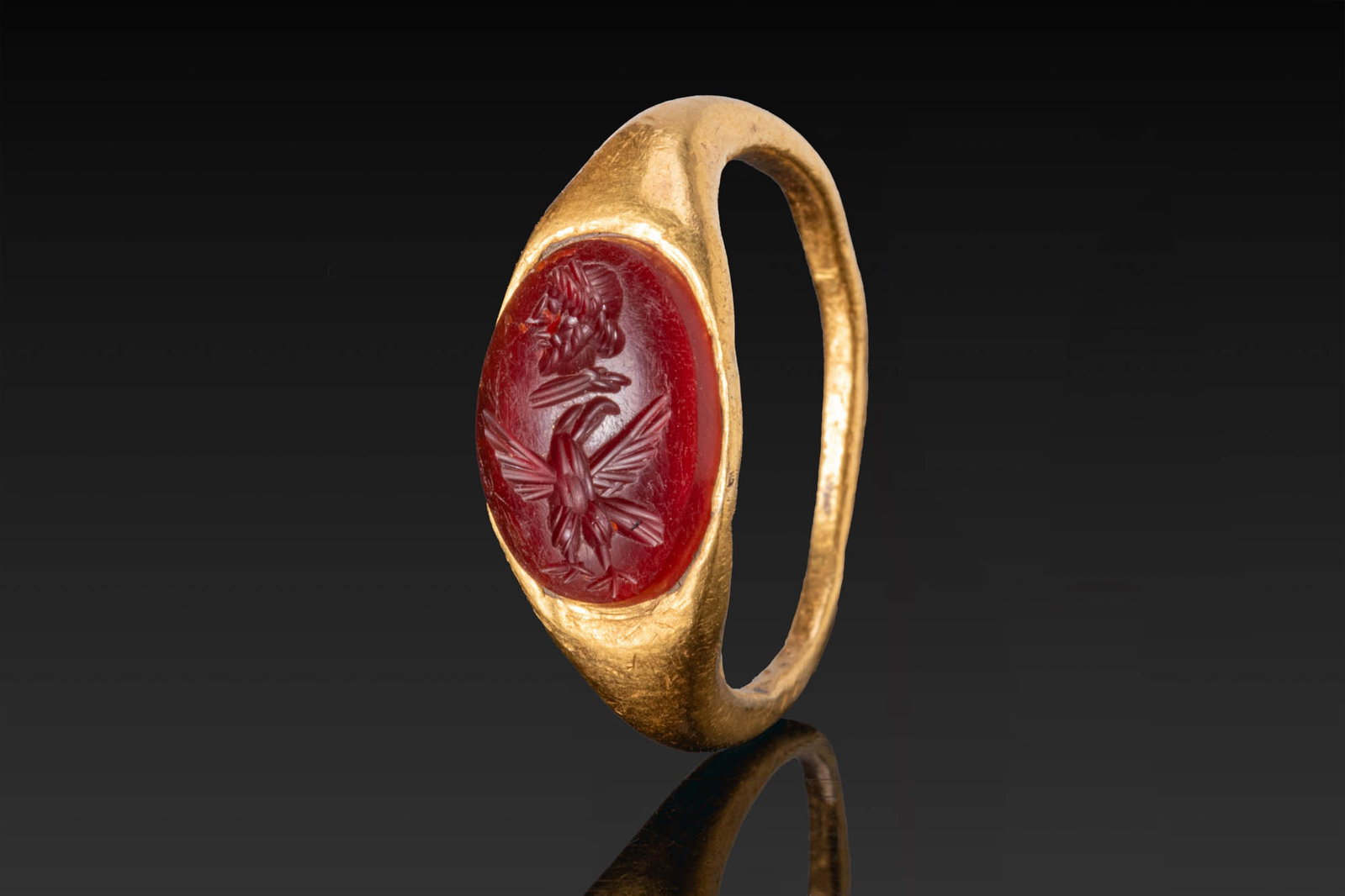 ROMAN GOLD RING WITH CARNELIAN INTAGLIO OF JUPITER AND EAGLE: Ca. AD 100 - 200. A gold ring with a slender hoop broadening to an oval bezel set with a carnelian intaglio engraved with the head of Jupiter in left profile above an eagle with spread wings. For simi