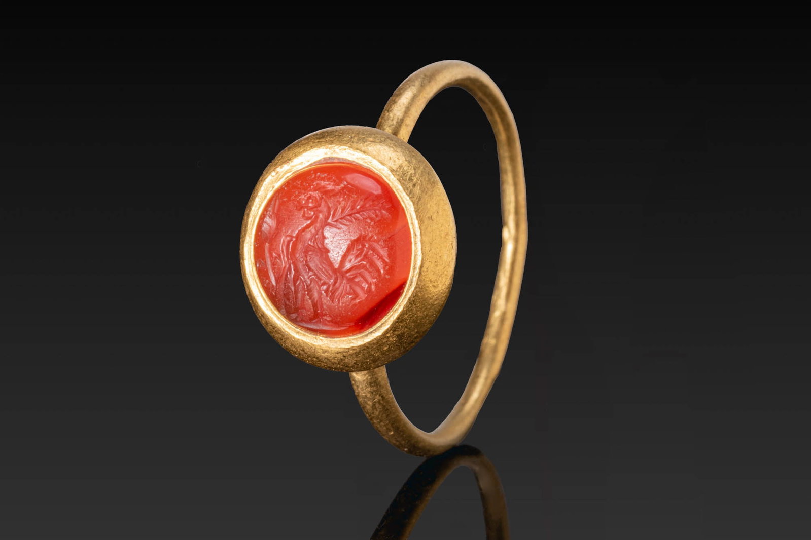 ROMAN GOLD RING WITH CARNELIAN INTAGLIO OF A COCKEREL: Ca. AD 300 - 500. A gold ring with a slender round-section hoop and a circular bezel set with a carnelian intaglio engraved with a standing cockerel in profile, with vegetal branches behind. For