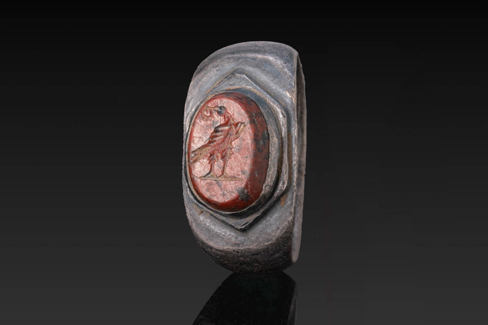 ROMAN RED STONE INTAGLIO OF AN EAGLE IN SILVER RING: Ca. AD 1 - 200. A red stone intaglio engraved with an eagle standing on a groundline and holding a laurel wreath in its beak, set in a possibly later silver ring with a broad, rounded hoop and a hexag