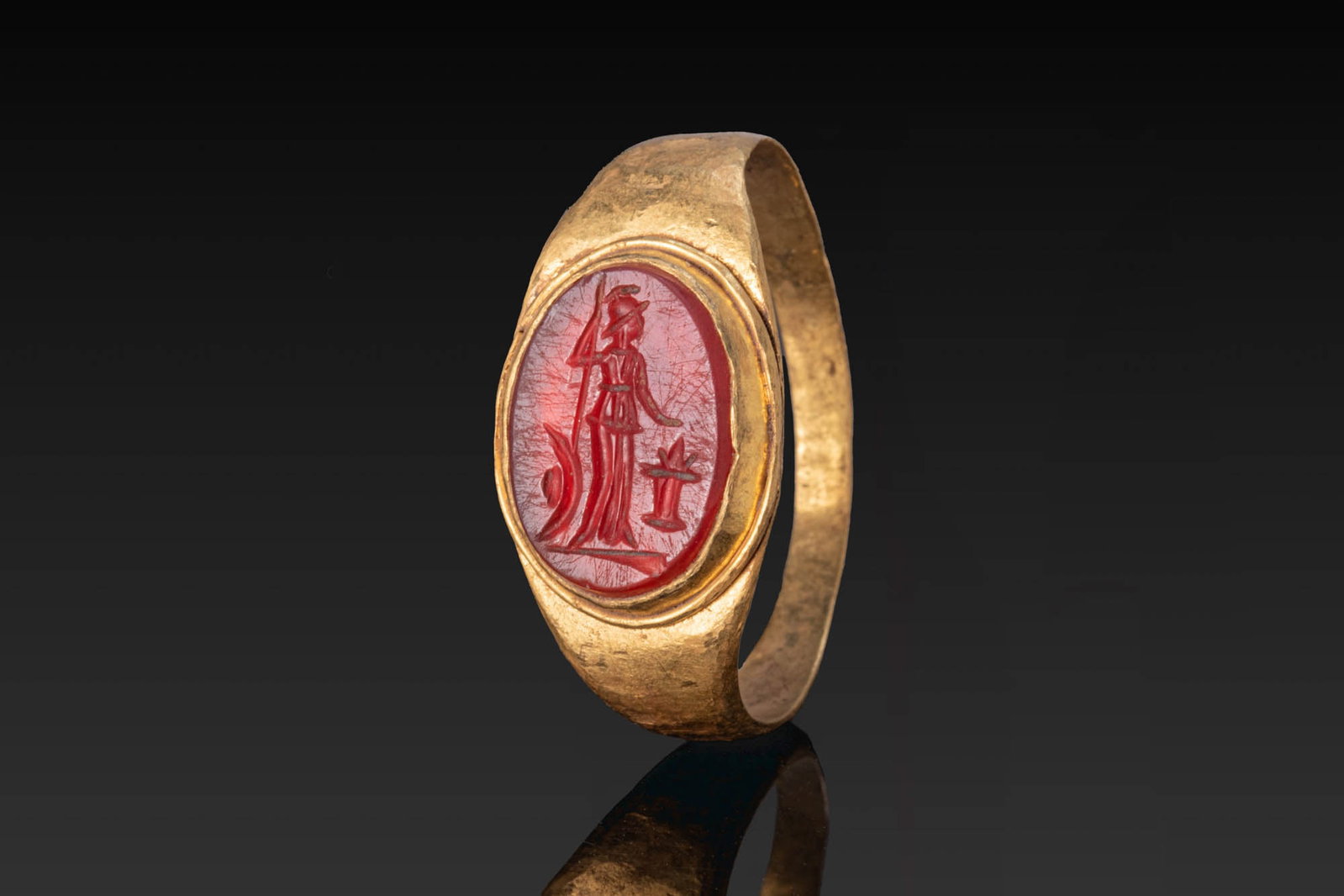 ROMAN GOLD RING WITH INTAGLIO OF MINERVA AND ALTAR: Ca. AD 1 - 300. A gold ring with a flat-section hoop, flaring shoulders, and an oval bezel set with a carnelian intaglio engraved with Minerva standing beside an altar, wearing a crested helmet and a 