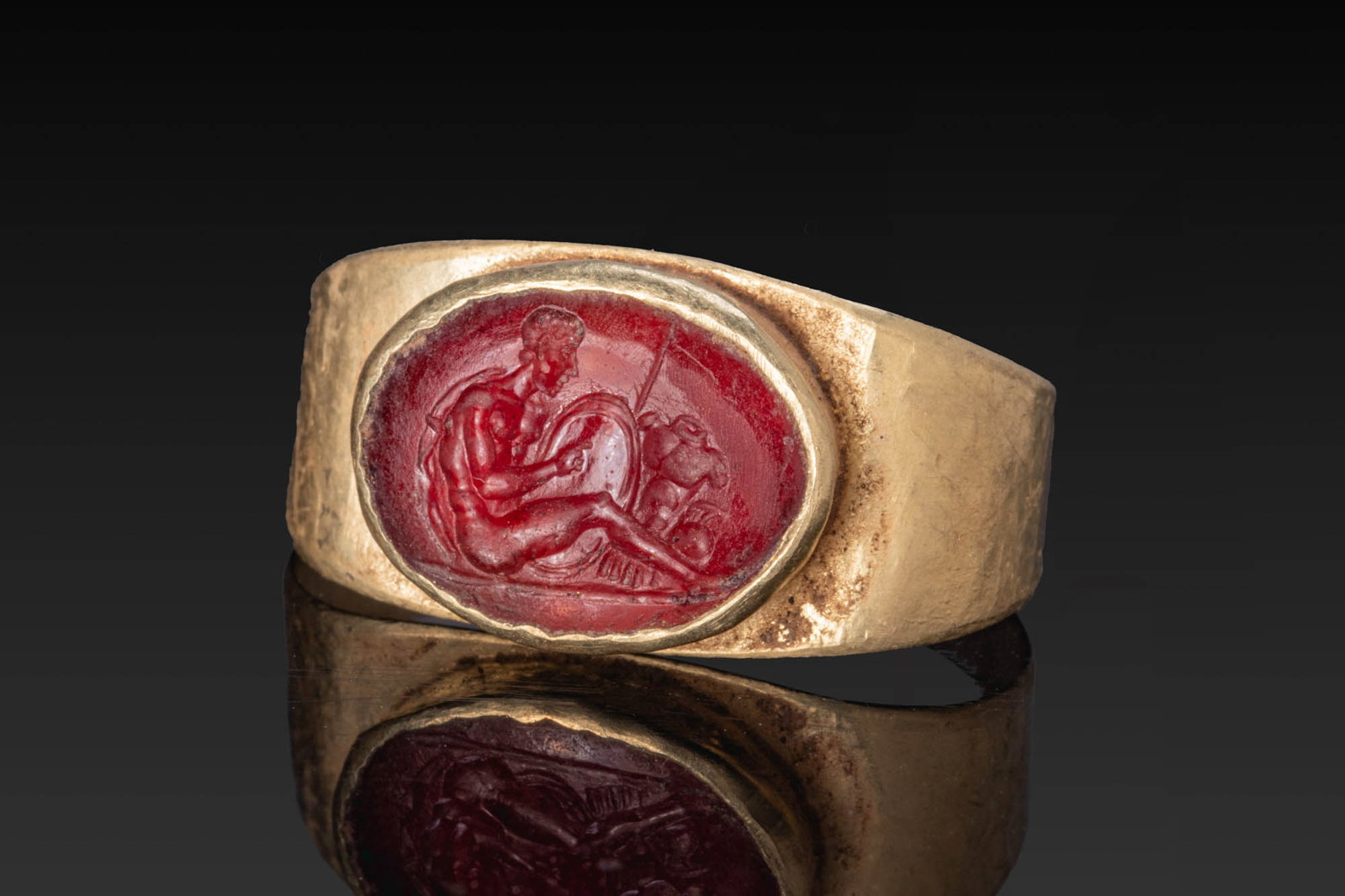 ROMAN GARNET INTAGLIO OF SEATED MARS WITH TROPHIES OF WAR IN GOLD RING: Ca. 100 BC - AD 100. A garnet intaglio engraved with Mars seated with a shield before him and trophies of war at his side, mounted in a possibly later gold ring with a broad tapering hoop and an oval 