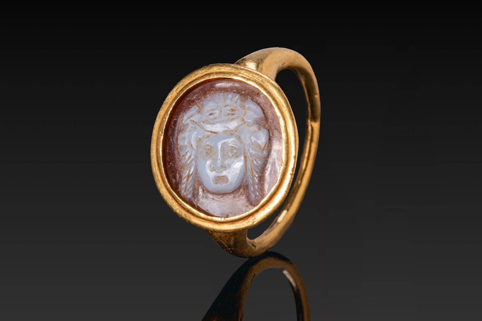 ROMAN AGATE CAMEO OF A THEATRICAL MASK IN GOLD RING: Ca. AD 1 - 300. A two-layered agate cameo carved with the frontal head of a theatrical mask, with staring eyes and stylised locks framing the face, mounted in a possibly later gold ring with a slender