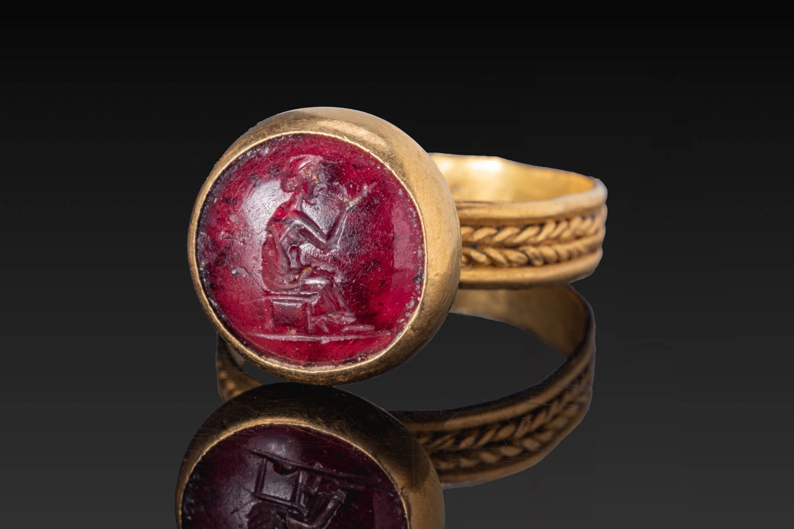 ROMAN RED GARNET INTAGLIO WITH SEATED FIGURE IN GOLD RING: Ca. AD 1 - 300. A red garnet intaglio engraved with a figure seated on a low stool on a groundline, turned to the right with the right arm raised, set in a possibly later gold ring. The hoop has a bra