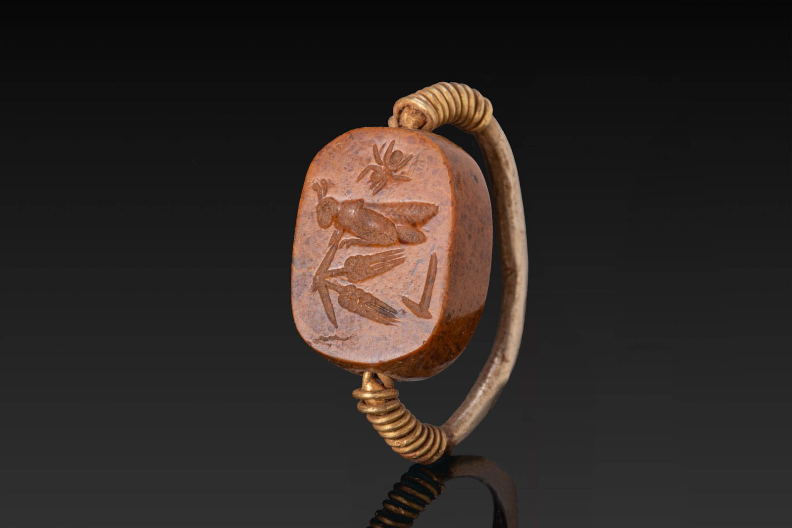 ROMANO-EGYPTIAN GOLD RING WITH BRECCIA SCARABOID WITH A SPIDER, GRASSHOPPER, AND WHEAT STALKS: Ca. 30 BC - AD 200. A gold ring with a rounded hoop, the breccia scaraboid bound at either end with coils, its upper face plain and convex, and its flat underside engraved with a spider above, a grass