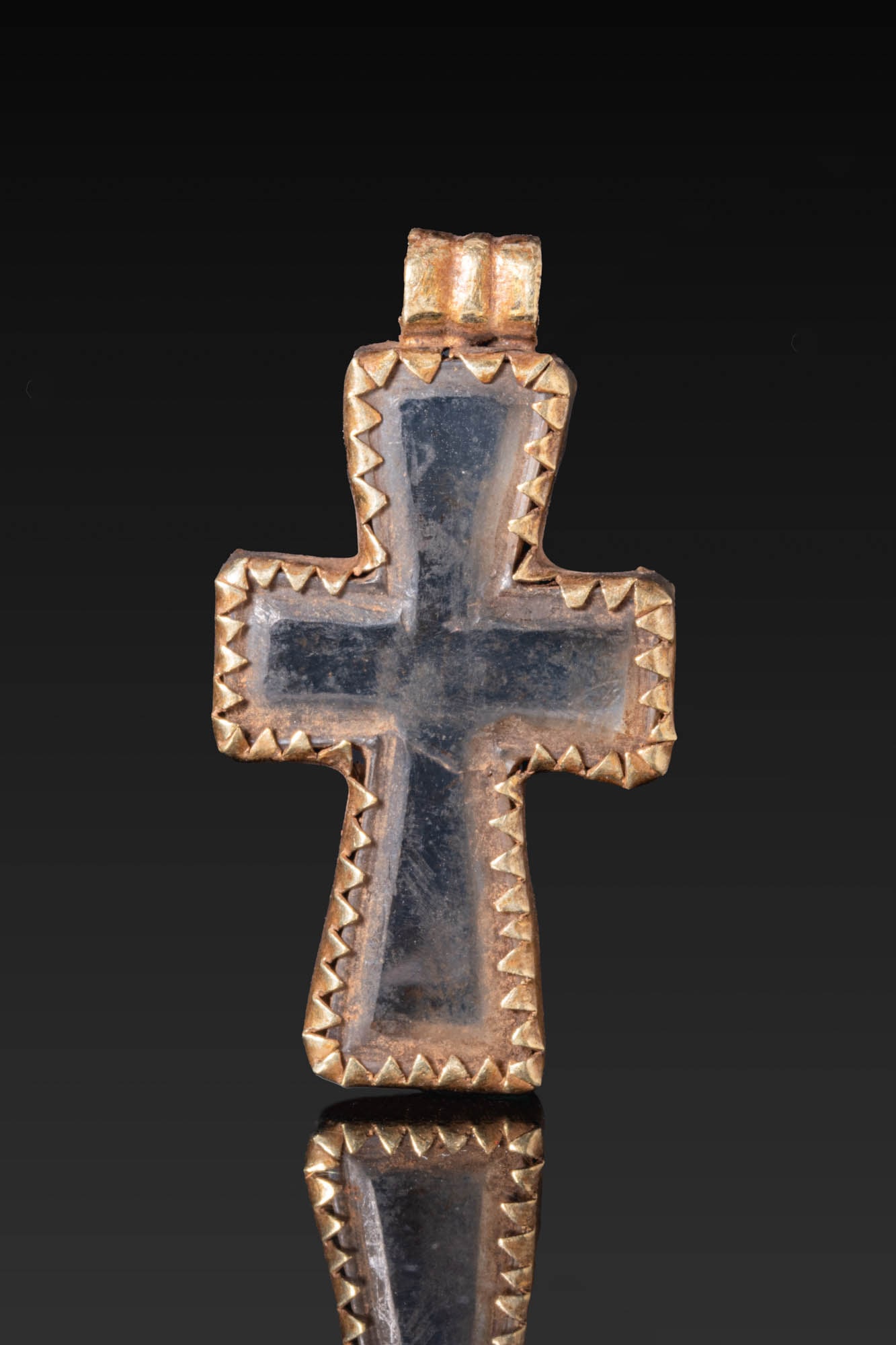 BYZANTINE GOLD PENDANT WITH GLASS CROSS: Ca. AD 1000 - 1200. A gold cruciform pendant with a cross-shaped glass inset in a serrated frame. The reverse is plain, and the pendant is fitted with a ribbed suspension loop above. Size: 39mm x