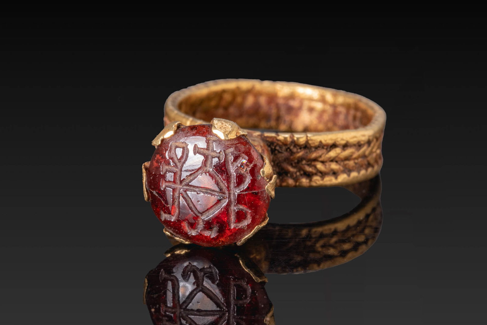 BYZANTINE GOLD RING WITH HIGH BEZEL AND GARNET INTAGLIO WITH MONOGRAM: Ca. AD 400 - 600. A gold ring with a broad hoop decorated with a braided band between raised borders, and a high conical bezel above a globular collar, set with a garnet intaglio engraved with a monog