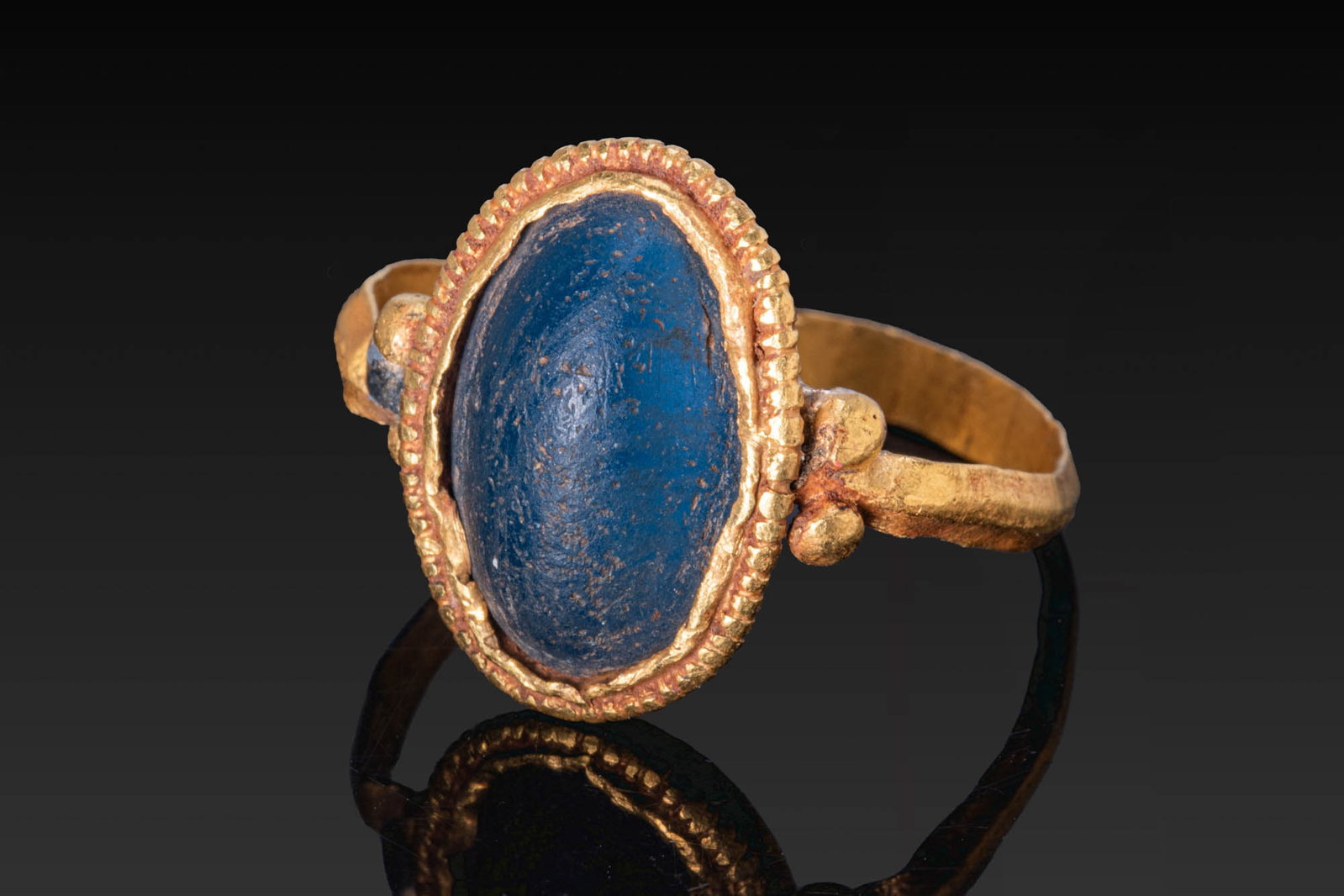 BYZANTINE GOLD RING WITH LARGE BLUE GLASS CABOCHON: Ca. AD 600. A gold ring with a slender carinated hoop and a large oval bezel set with a blue glass cabochon within a beaded border. Pairs of small globules flank the setting. For a similar cabochon, s