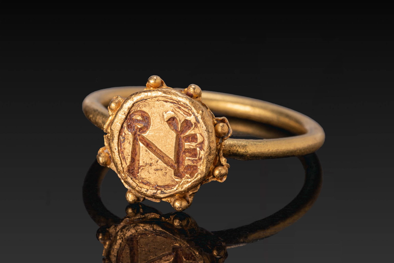 BYZANTINE GOLD RING WITH MONOGRAM: Ca. AD 400 - 600. A gold ring with a plain round-section hoop and a circular bezel framed by eight applied pellets, the underside formed as a fluted setting, the face engraved with a monogram. For sim