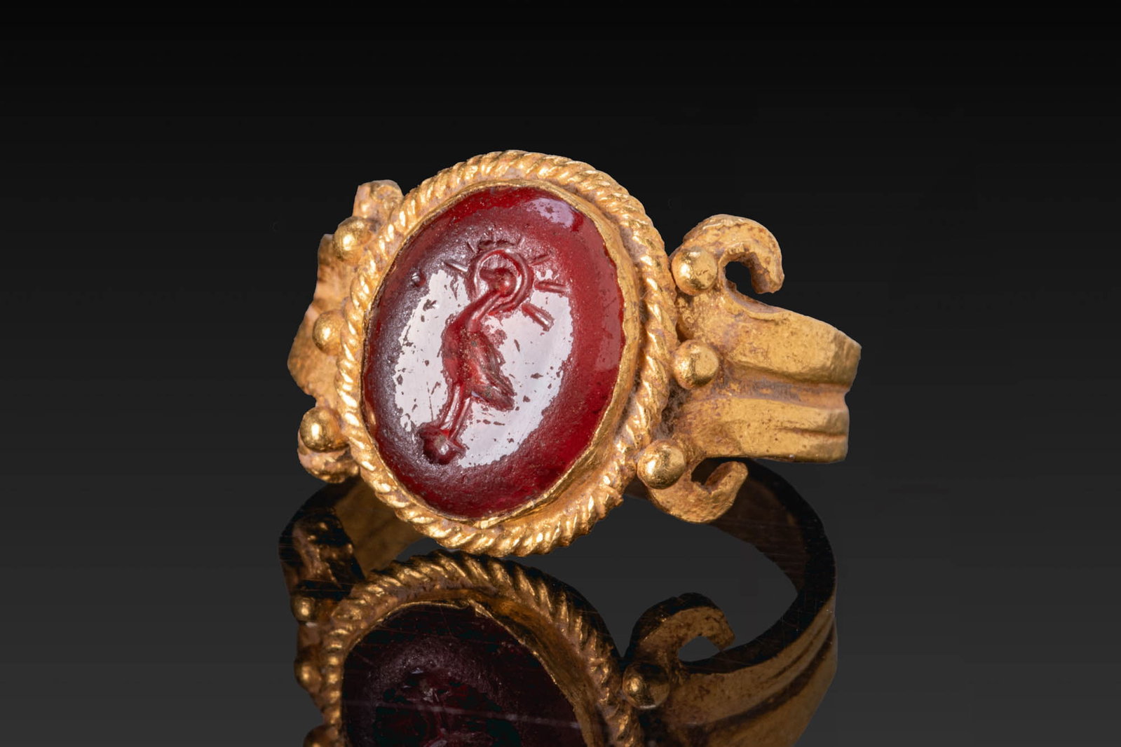 BYZANTINE GOLD RING WITH RED GARNET INTAGLIO OF A CROWNED CRANE: Ca. AD 300. A gold ring with a flat-section hoop with a central groove, bifurcated shoulders with scrolls and three gold pellets, and an oval bezel cell with a twisted wire border, set with a convex r