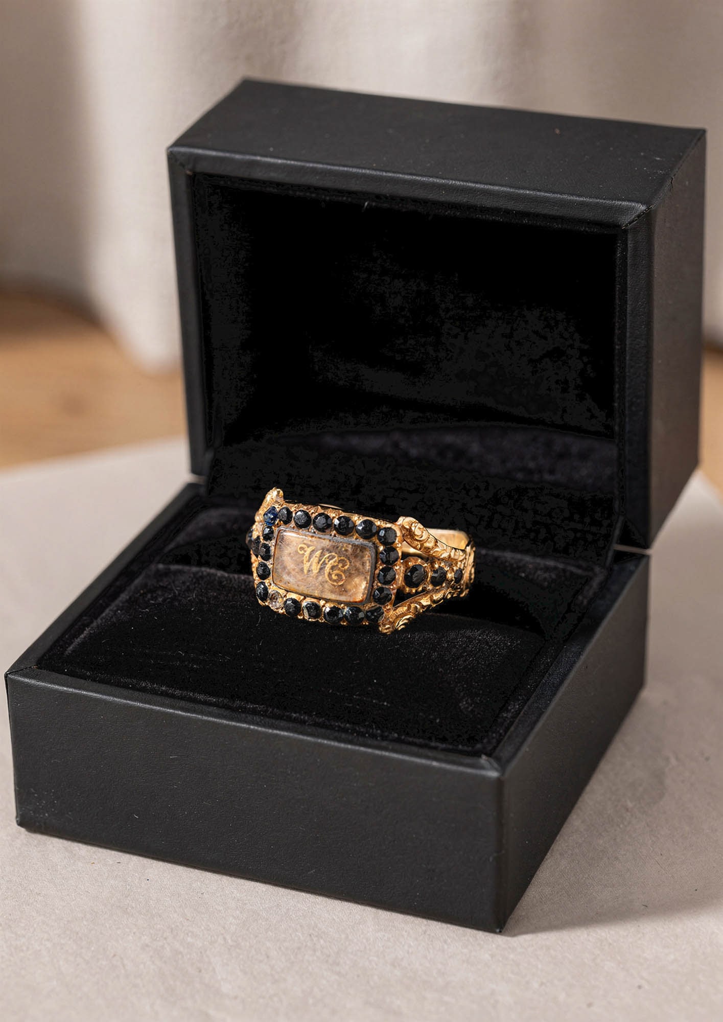GEORGIAN OPENWORK GOLD RING WITH GLASS PLAQUE, SAPPHIRES, AND INITIALS "WC": Ca. AD 1820. A gold openwork mourning ring with a rectangular glass panel enclosing hairwork and the initials "WC" at the centre, within a border of round cut sapphires. The bifurcated shoulders are c