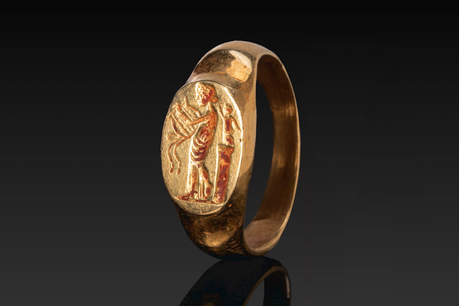 BACTRIAN SOLID GOLD RING WITH APOLLO CITHAROEDUS AND STATUE ON PILLAR: Ca. AD 1 - 300. A solid gold ring with a broad hoop, recessed shoulders, and an oval bezel engraved with Apollo Citharoedus standing in profile and holding a cithara, with a small statue on a column b