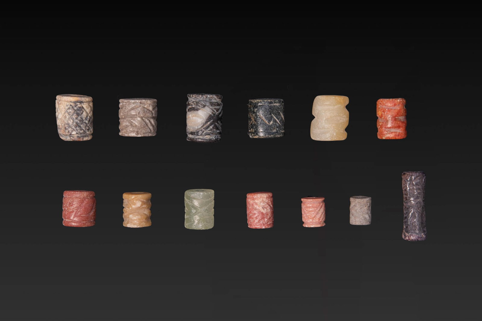 LARGE COLLECTION OF JEMDET NASR STONE CYLINDER SEALS WITH GEOMETRIC MOTIFS: Ca. 3000 BC. A collection of 13 stone cylinder seals of pale, black, green, yellow, and red stone, each carved with geometric motifs, including cross-hatched fields, zigzags, and chevrons. Size: 7-16m