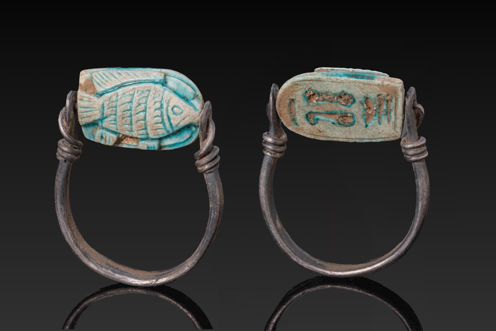 EGYPTIAN GLAZED STEATITE FISH SCARABOID IN SILVER SWIVEL RING: New Kingdom, Ca. 1550 - 1069 BC. A turquoise-glazed steatite Tilapia fish scaraboid with an incised eye, hatched fins, and a fanned tail. The flat underside is engraved with a hieroglyphic inscription