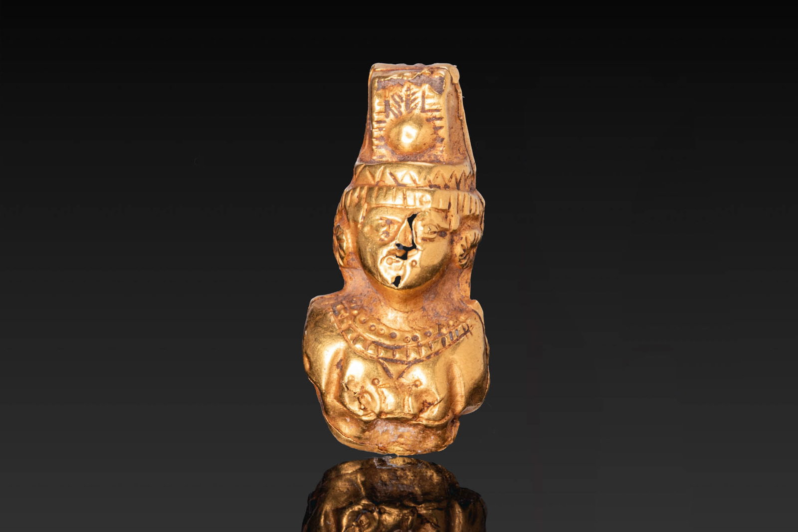 EGYPTIAN GOLD PENDANT OF AMUN: Ptolemaic Period, Ca. 332 - 30 BC. A gold pendant in the form of the bust of Amun, with a rounded face and striated hair, surmounted by a tall crown centred with a sun disc. The chest is adorned with 