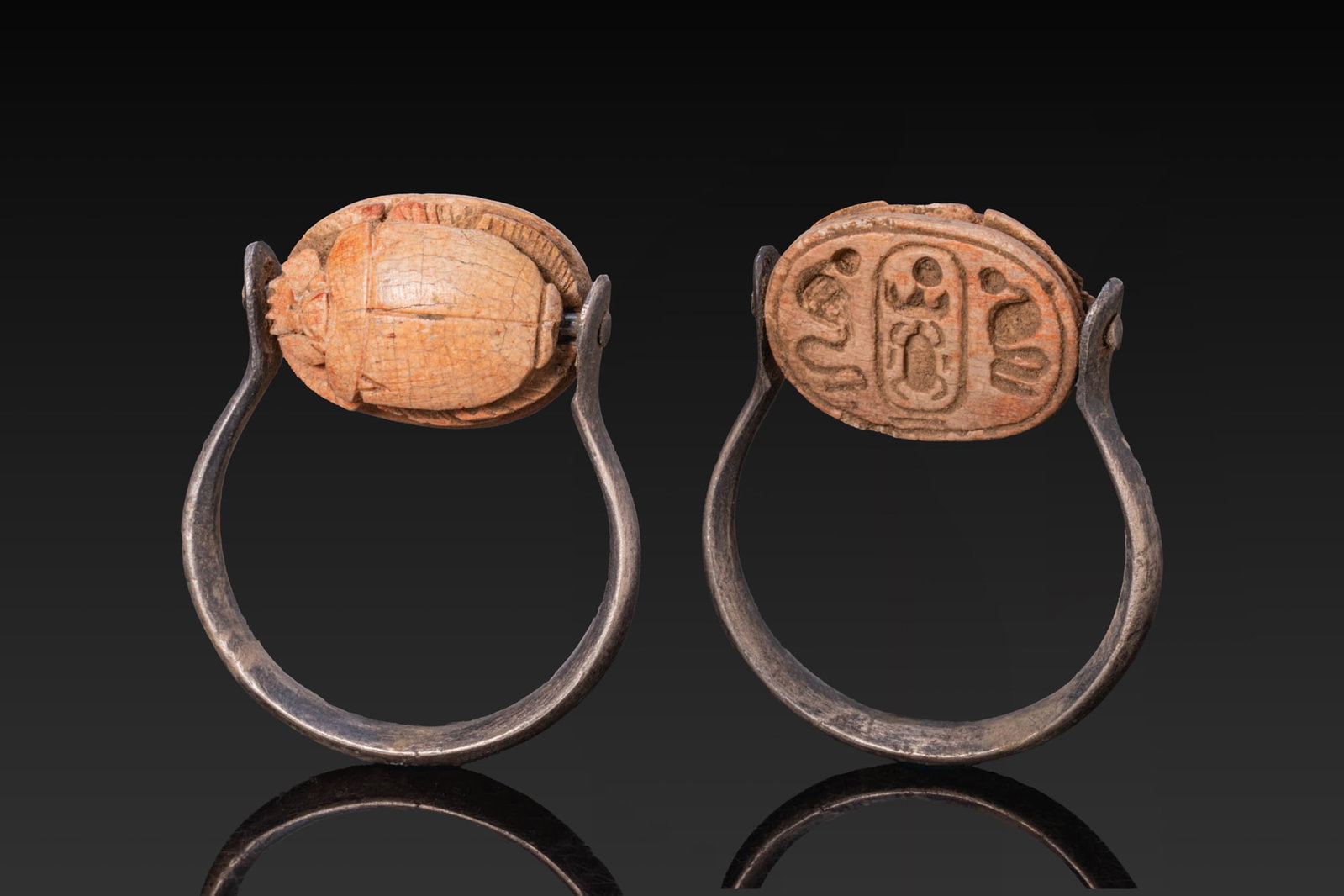 EGYPTIAN STEATITE SCARAB WITH CARTOUCHE OF THUTMOSE III BETWEEN URAEI IN SILVER SWIVEL RING: New Kingdom, Ca. 1550 - 1069 BC. A steatite scarab with a domed back, a single line marking the suture and pronotum, and v-marks at the upper outer edges of the elytra. The legs are incised and natura