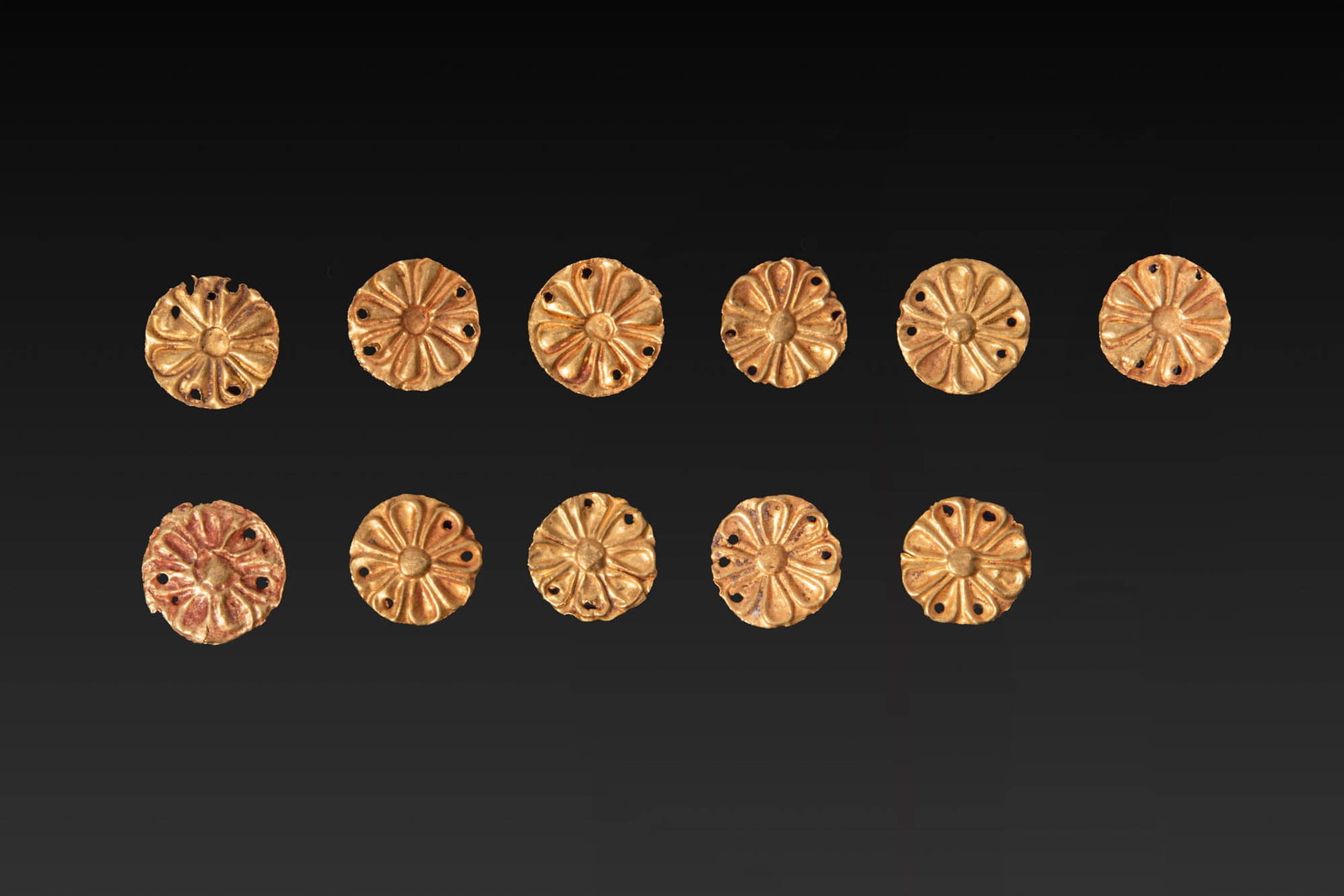COLLECTION OF GREEK GOLD ROSETTE APPLIQUES: Ca. 600 - 200 BC. A collection of 11 gold rosette appliques, each with a raised central boss surrounded by radiating almond-shaped petals in repousse. Small perforations are pierced around the perimet