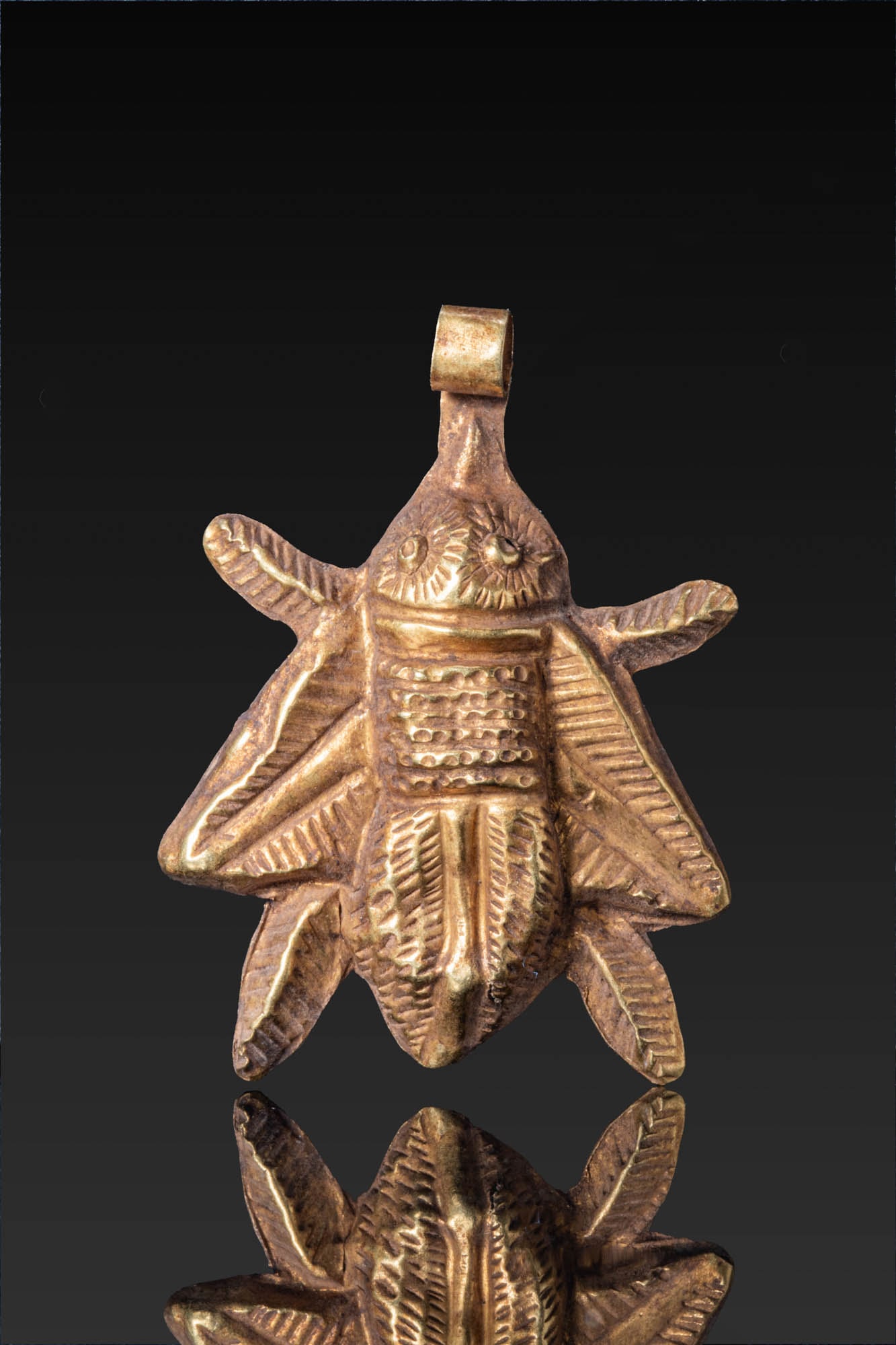 GREEK HIGH KARAT GOLD FLY PENDANT: Ca. 600 - 400 BC. A gold fly pendant formed from a thin hammered sheet with repousse modelling of the insect, with a rounded head with pellet eyes, a broad thorax flanked by folded wings, and a taperi