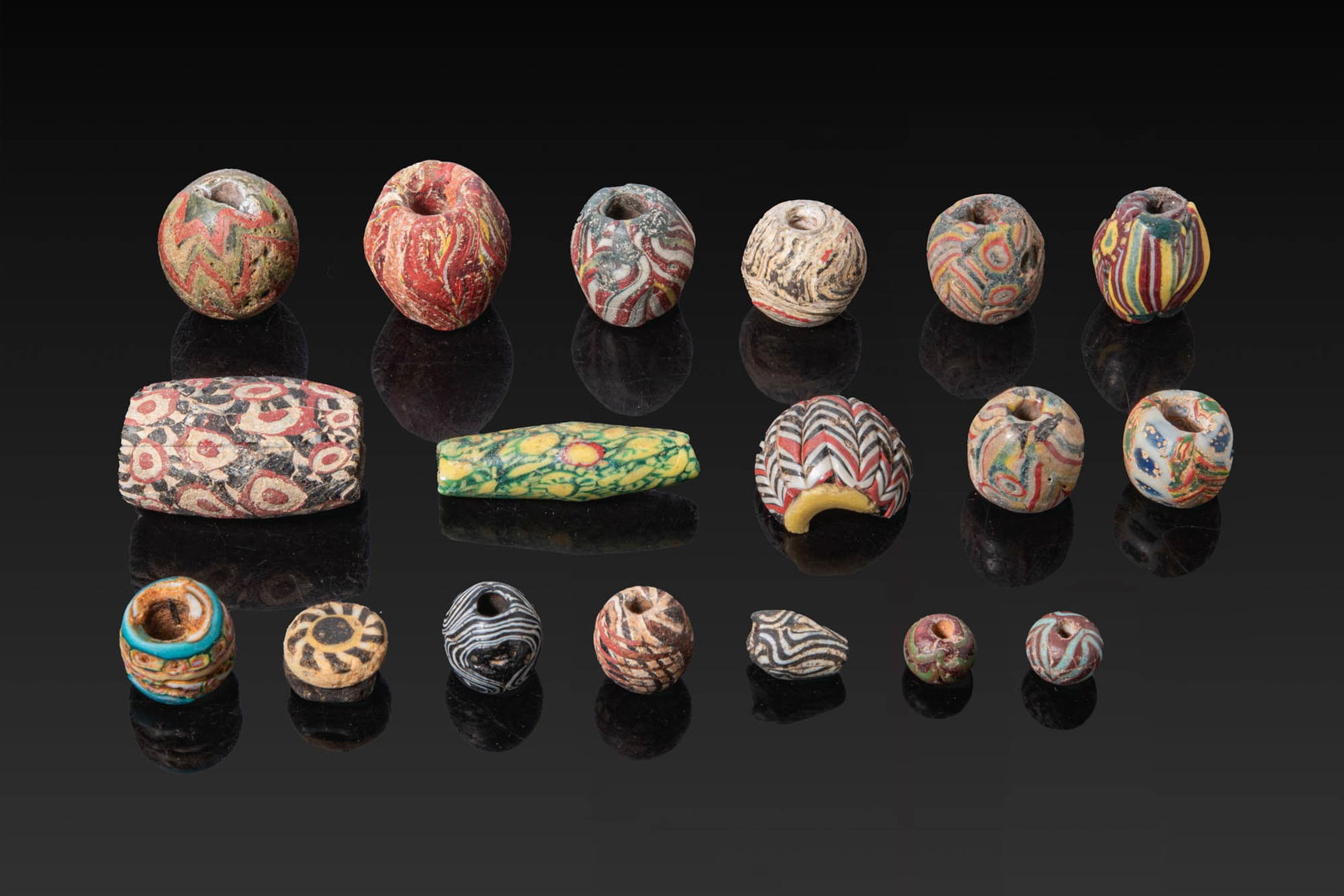 COLLECTION OF GREEK AND ROMAN GLASS BEADS: Ca. 500 BC - AD 200. A collection of 18 glass beads of rounded, cylindrical, and barrel shapes. The group includes beads with combed and trailed bichrome decoration, mosaic canes arranged in geometric