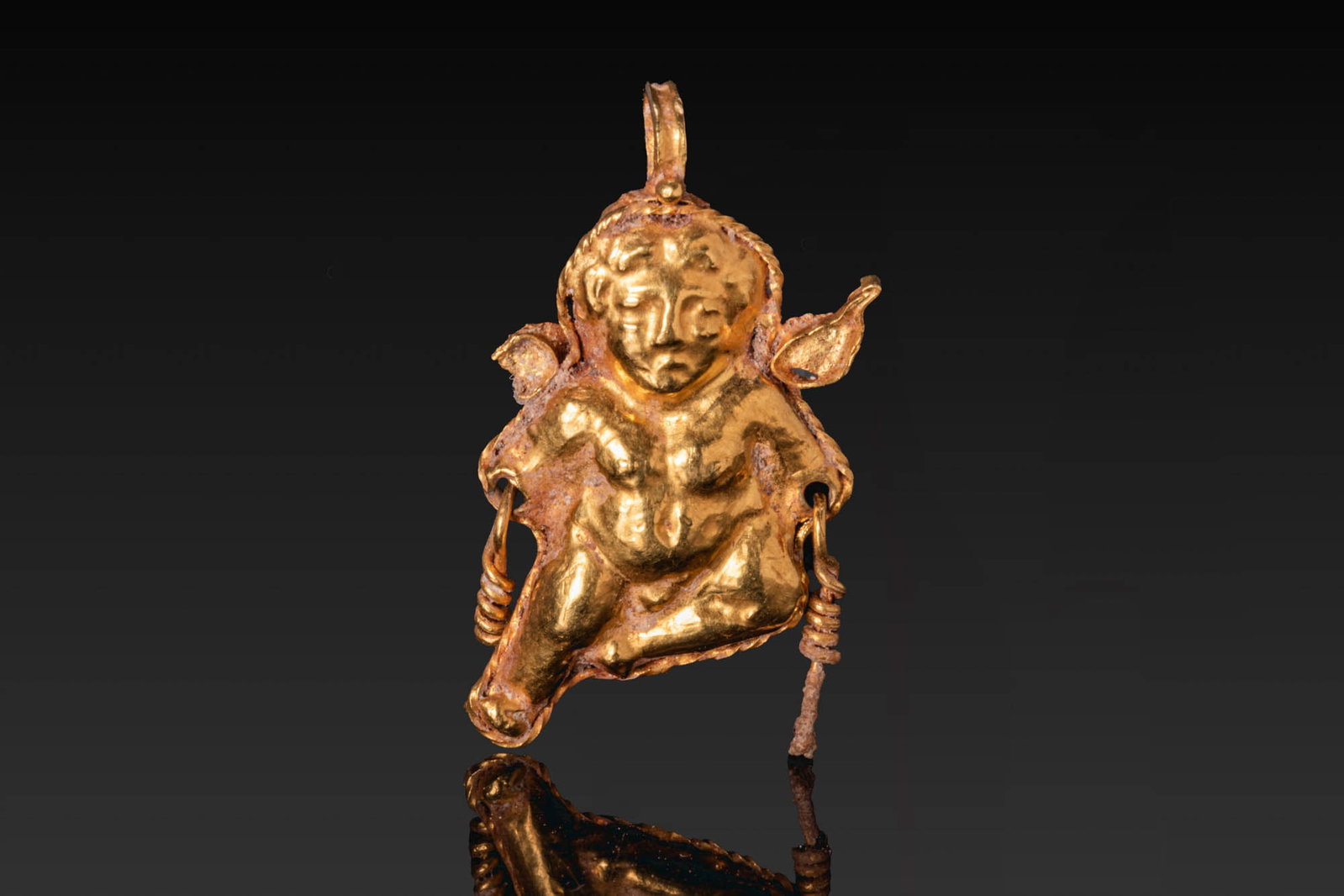HELLENISTIC GOLD EROS PENDANT: Ca. 300 - 100 BC. A gold pendant of Eros seated nude, with his left leg bent and his right leg extended, with a suspension loop above and twisted wire attachments at the sides. For similar see: The Br