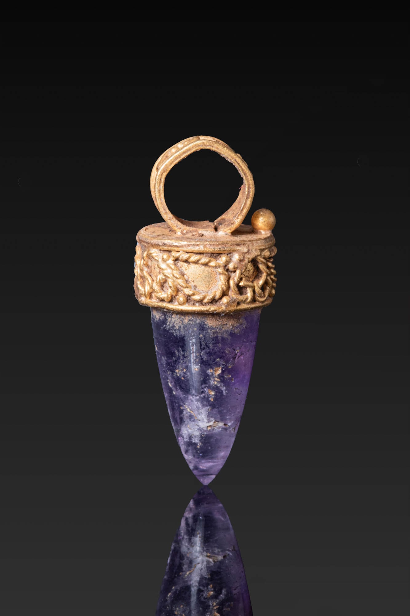 HELLENISTIC HIGH KARAT GOLD PENDANT WITH AMETHYST: Ca. 300 - 100 BC. A gold pendant set with a tapered amethyst drop in a circular mount with a plain upper plate, the collar and suspension loop decorated with granulation and twisted wire scrolls. Size